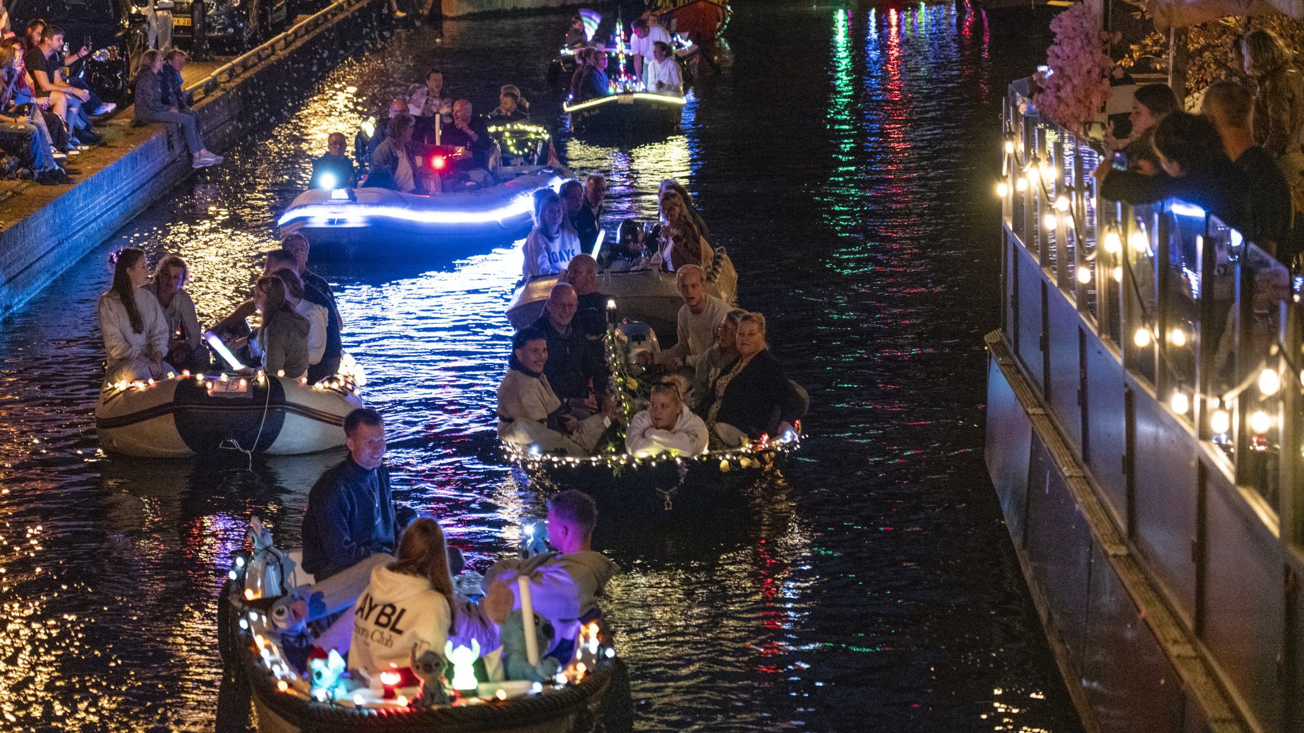 Kleine boten met mensen varen 's avonds over een verlichte gracht, omgeven door toeschouwers langs de kade.