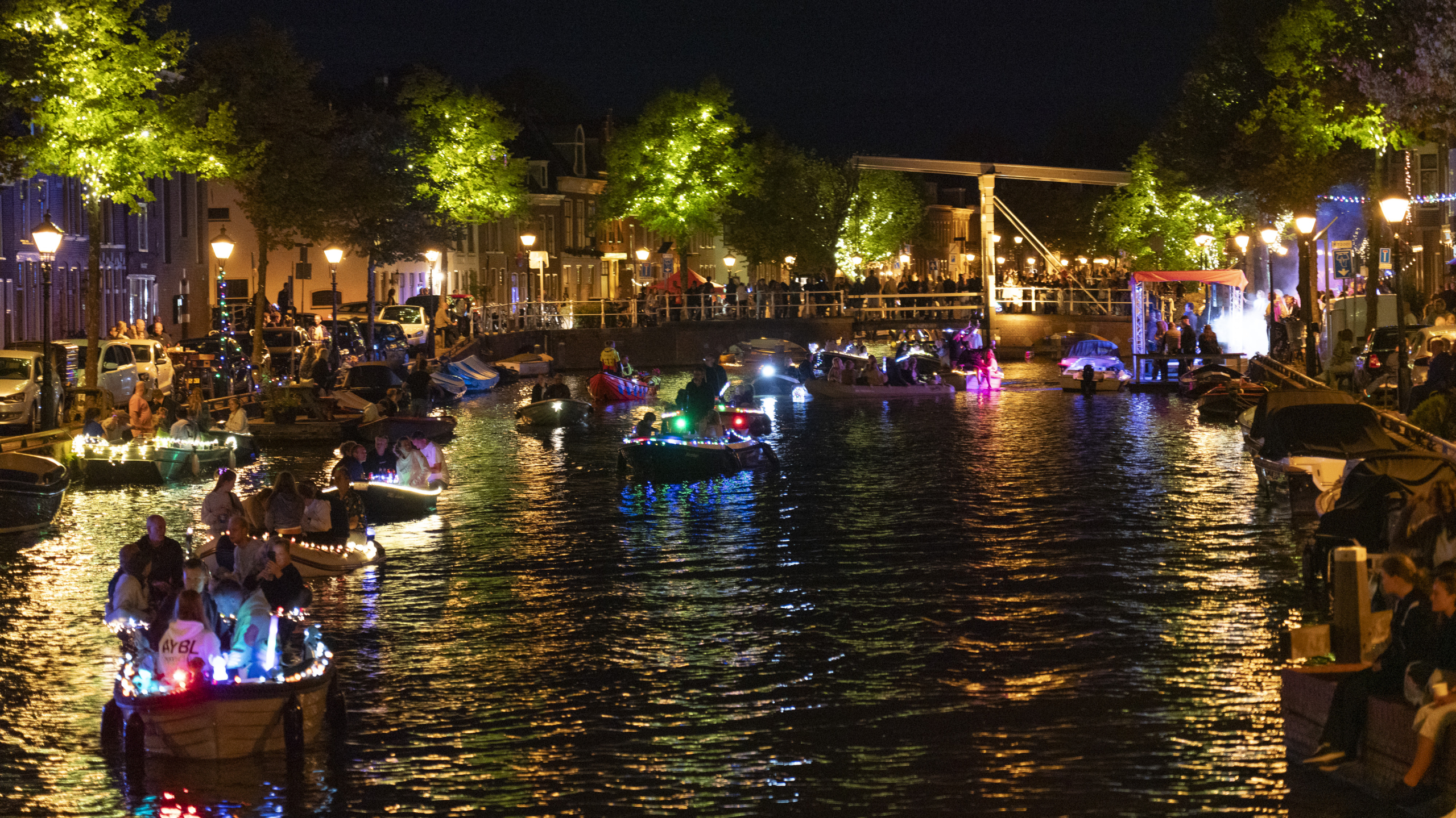 Avondparade van boten versierd met kleurrijke lichten op een verlichte gracht, met feestende mensen langs de waterkant.