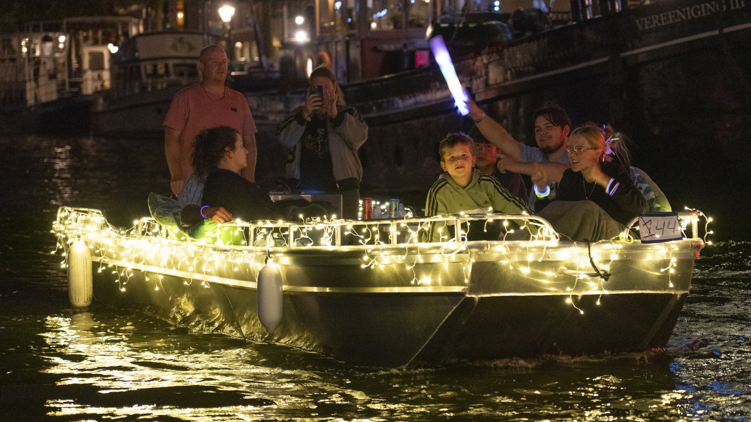 Een groep mensen vaart in een versierde boot met feestverlichting over het verlichte water, terwijl een persoon een lichtgevende staaf omhoog houdt.