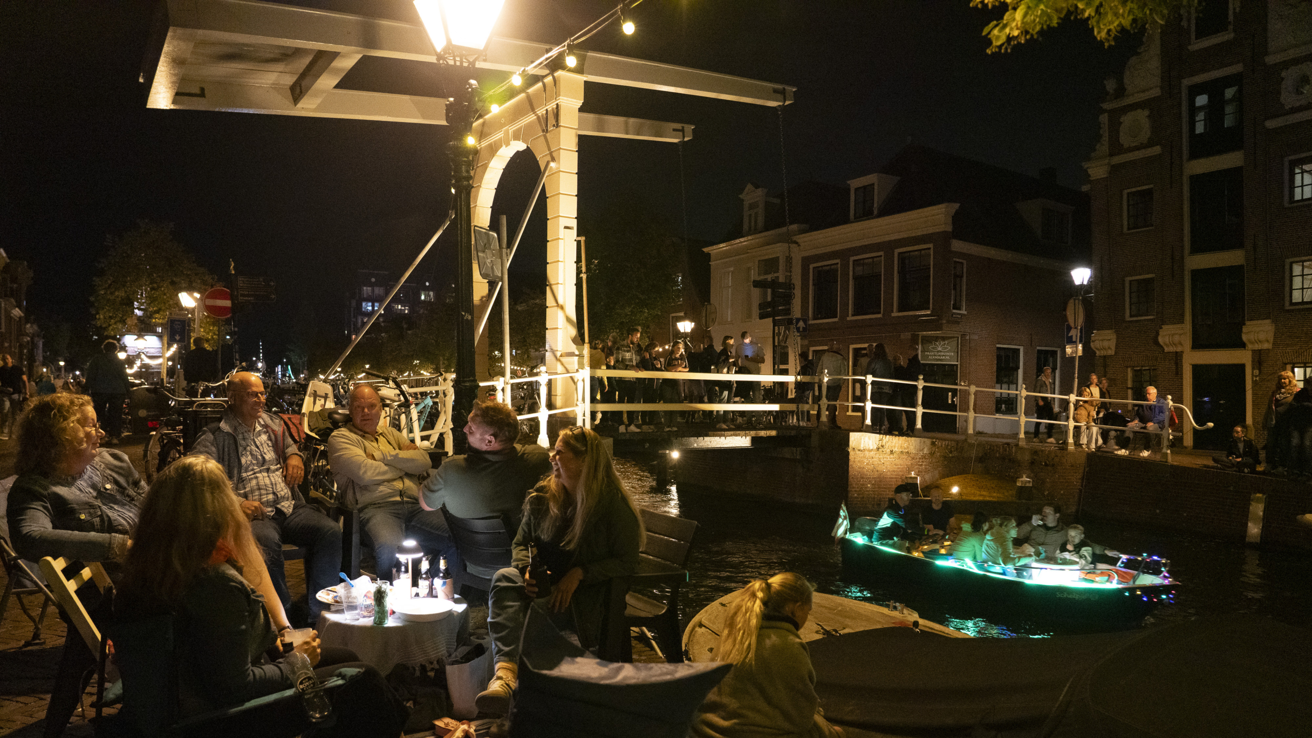 Een groep mensen zit 's avonds op een terras onder een straatlantaarn bij een verlichte brug in Amsterdam, met een bootje versierd met lichtjes in de gracht eronder.