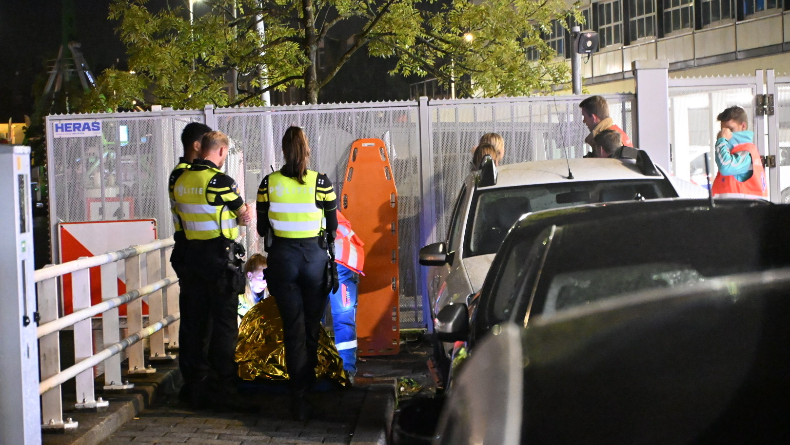 Politieagenten in uniform staan bij een persoon gedekt met een thermodeken op straat naast een geparkeerde auto en wegafzettingen in de avond.