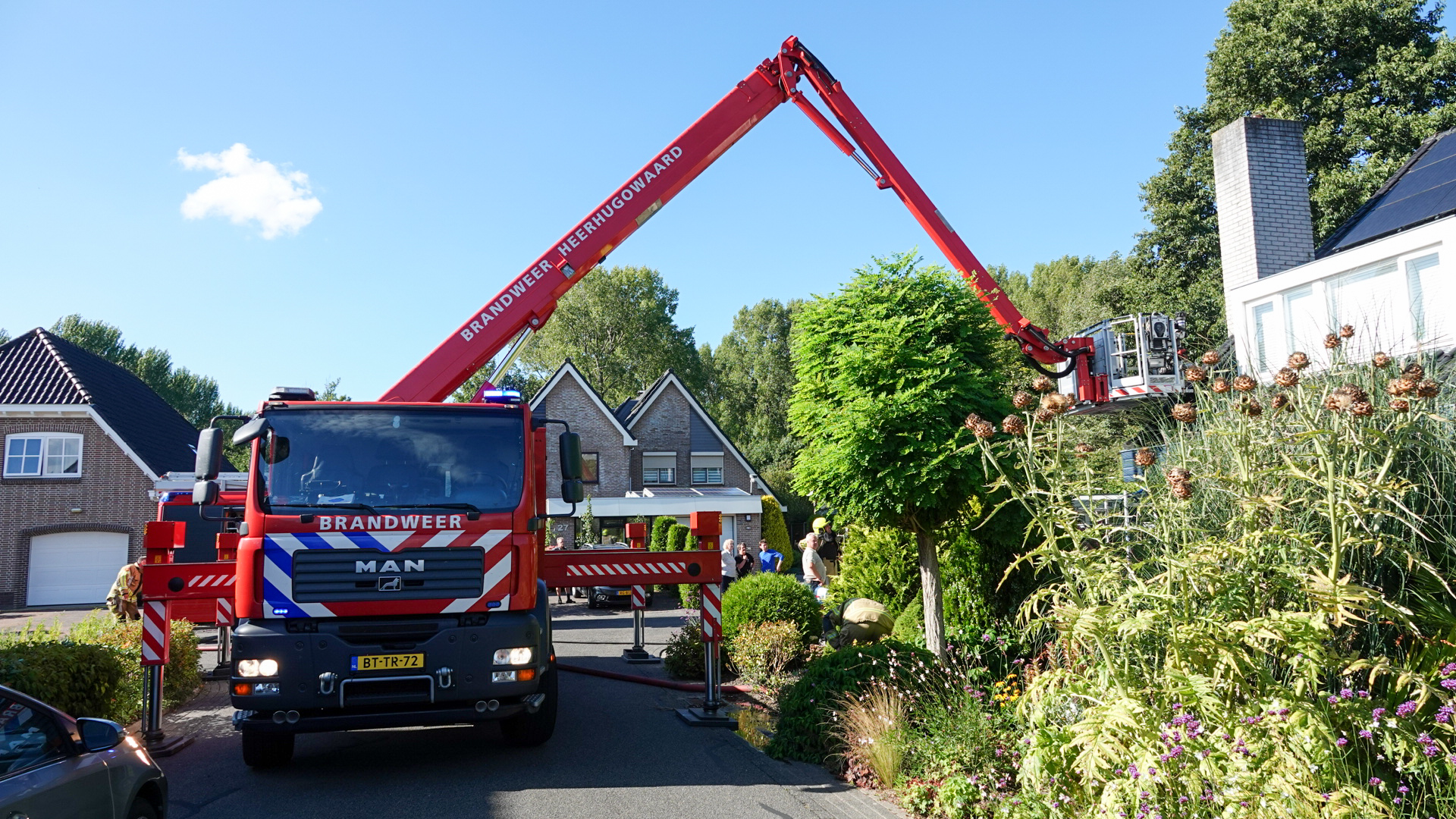 Een brandweerwagen van Brandweer Heerhugowaard staat met een uitgeschoven ladder in een woonwijk, omringd door huizen en groene bomen en struiken.