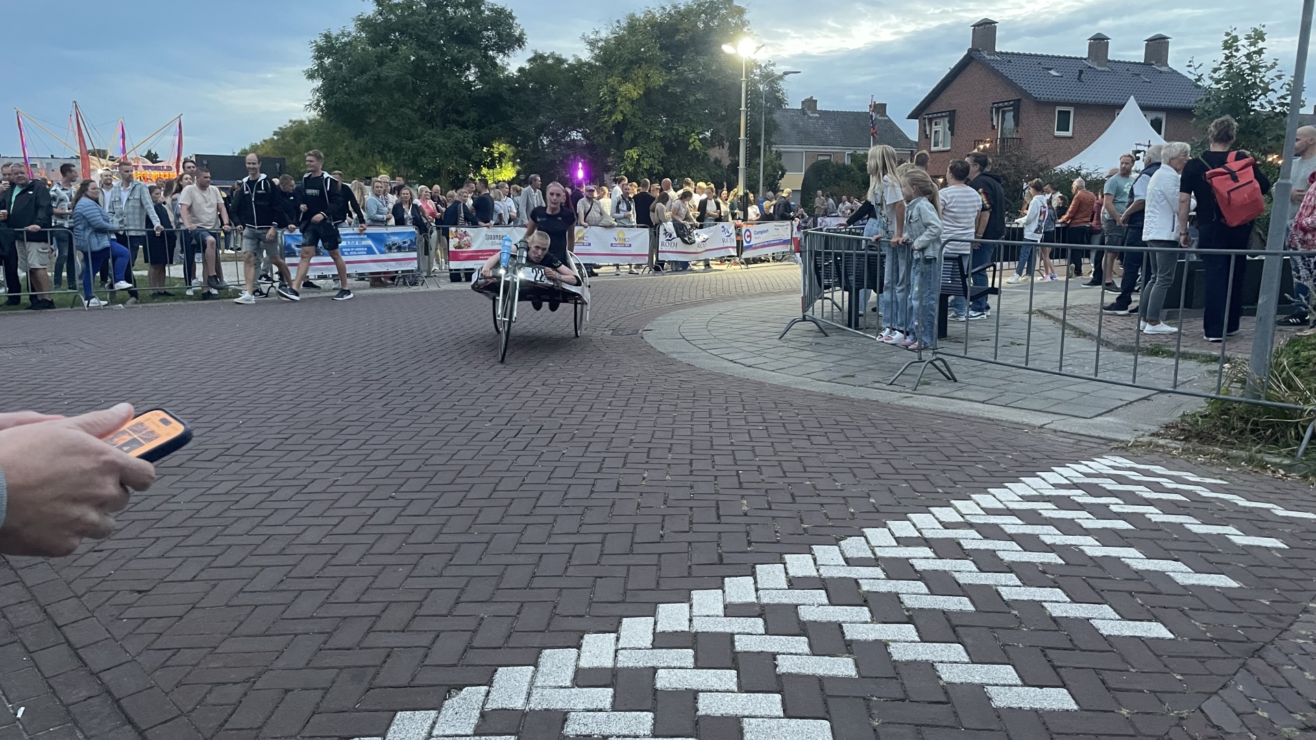 Mensen kijken toe hoe een ligfietsrijder met snelheid door een bocht rijdt tijdens een evenement, met toeschouwers die achter hekken staan langs de route en een kermisattractie zichtbaar op de achtergrond.