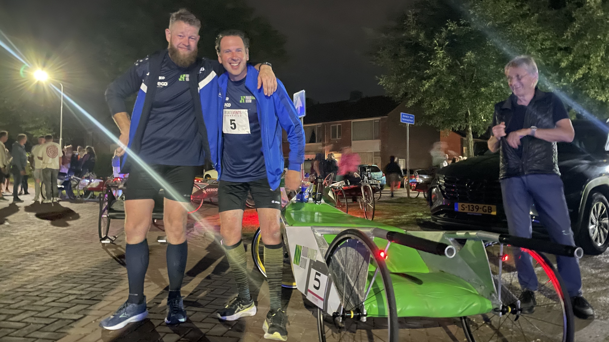 Twee mannen in sportkleding staan naast een driewielige kart, omringd door nachtelijke verlichting, met een oudere man die toekijkt.