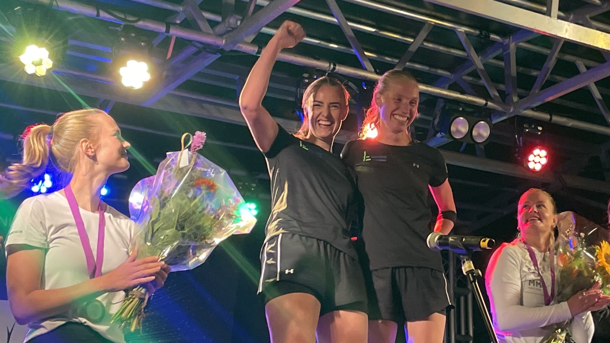 Vijf sporters staan op een podium met bloemen, terwijl ze in sportkleding onder felgekleurde lichten worden gehuldigd.