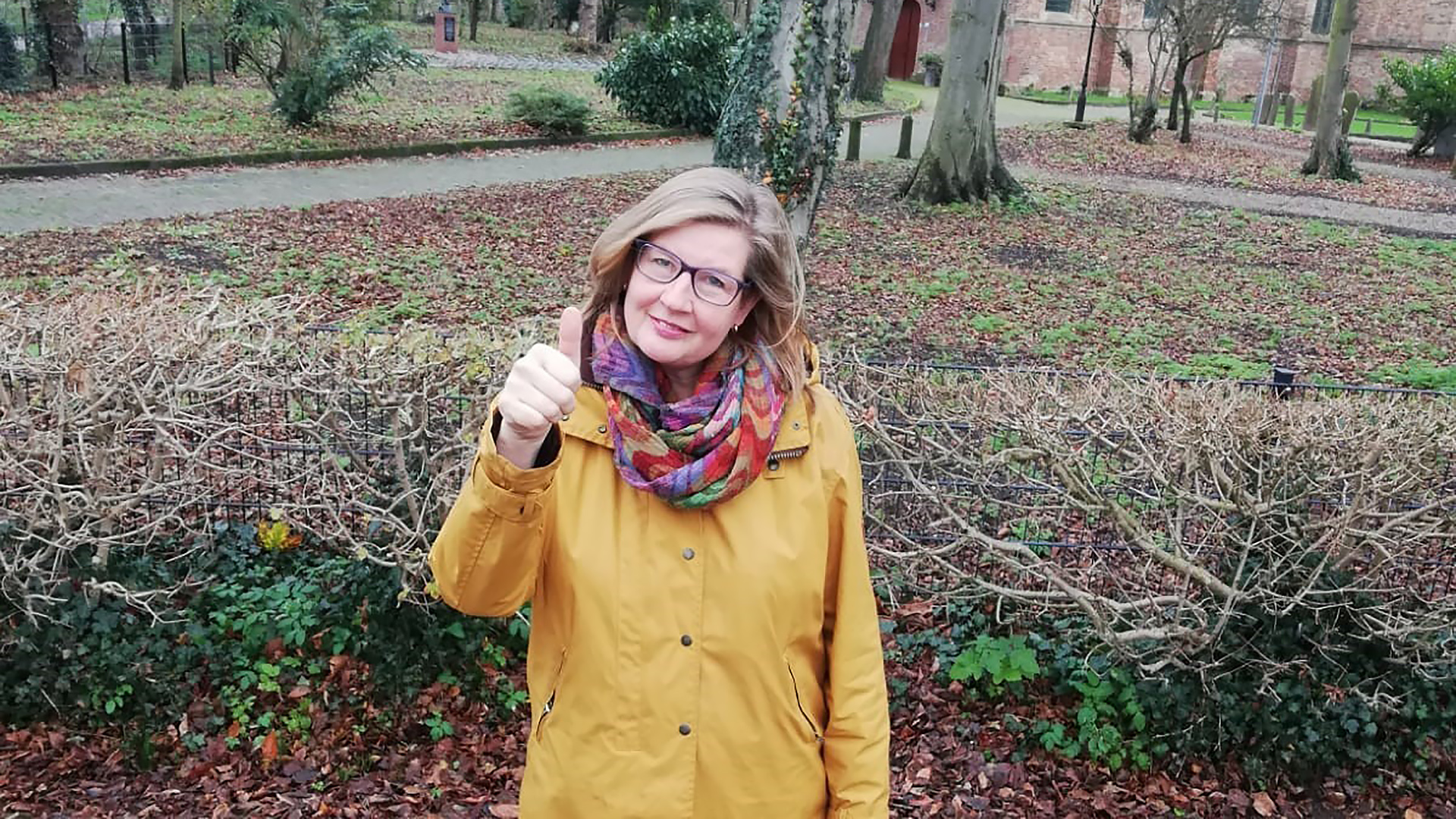 Vrouw met een gele jas en kleurrijke sjaal staat buiten, voor kale heggen en bomen, met een duim omhoog.