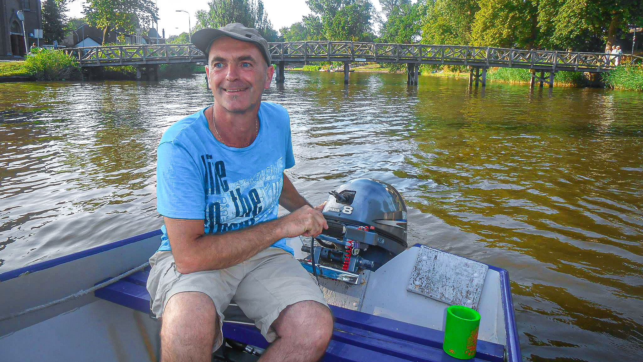 Man met hoed vaart een kleine motorboot op een rivier met een houten brug en groene bomen op de achtergrond.