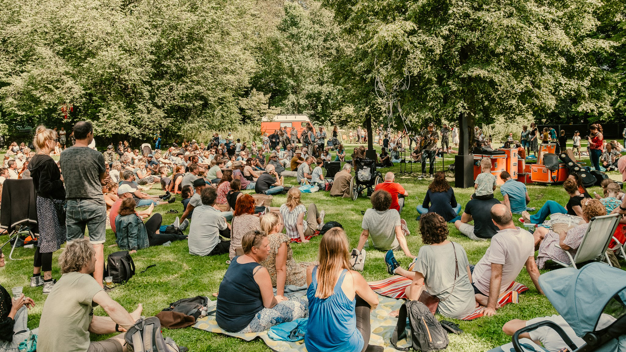 Een grote groep mensen zit en staat op het gras in een park, luisterend naar een live muziekoptreden onder de bomen.