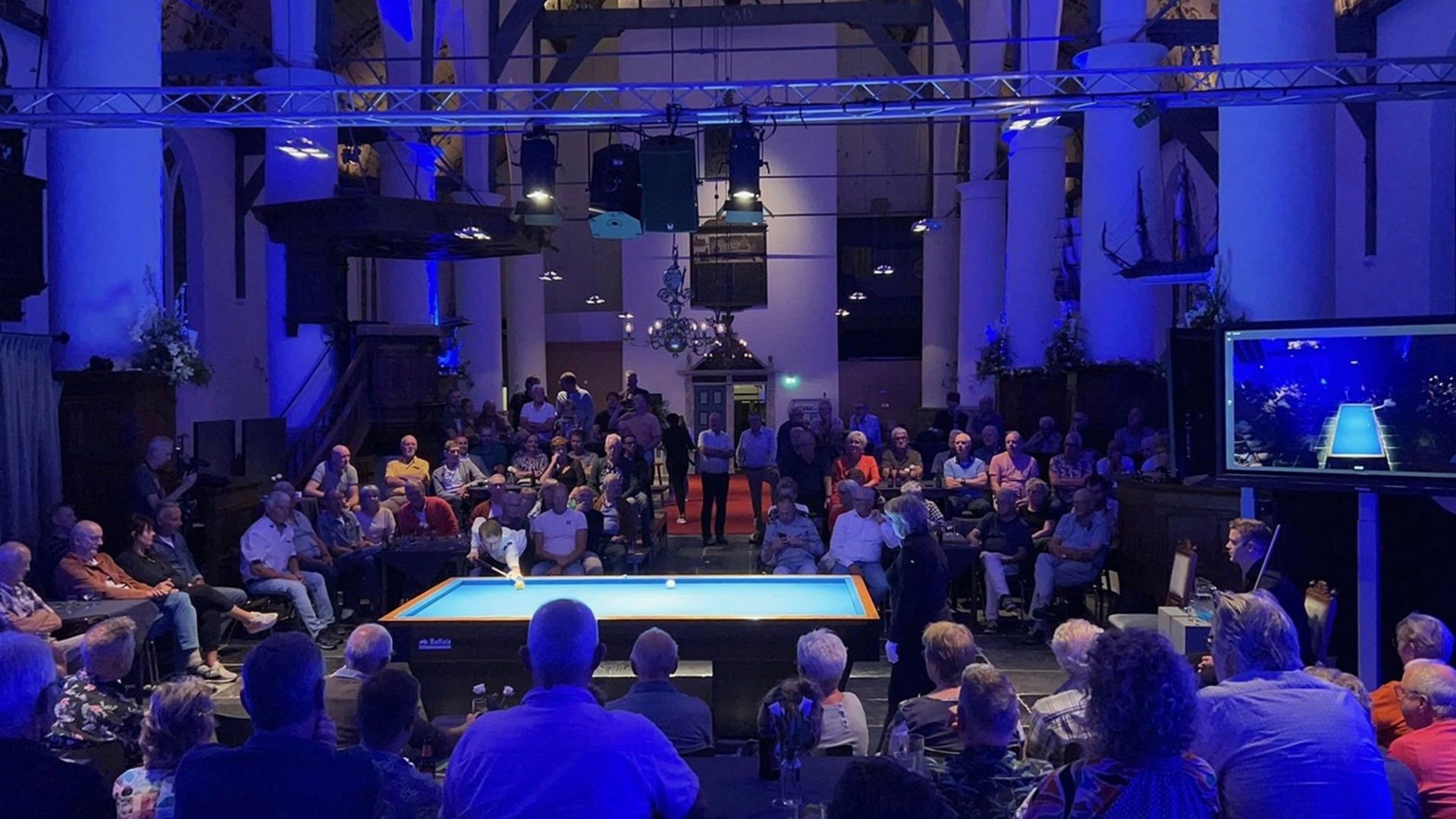 Biljartwedstrijd in een kerk met toeschouwers rondom een biljarttafel, sfeervol verlicht met blauwe lampen.