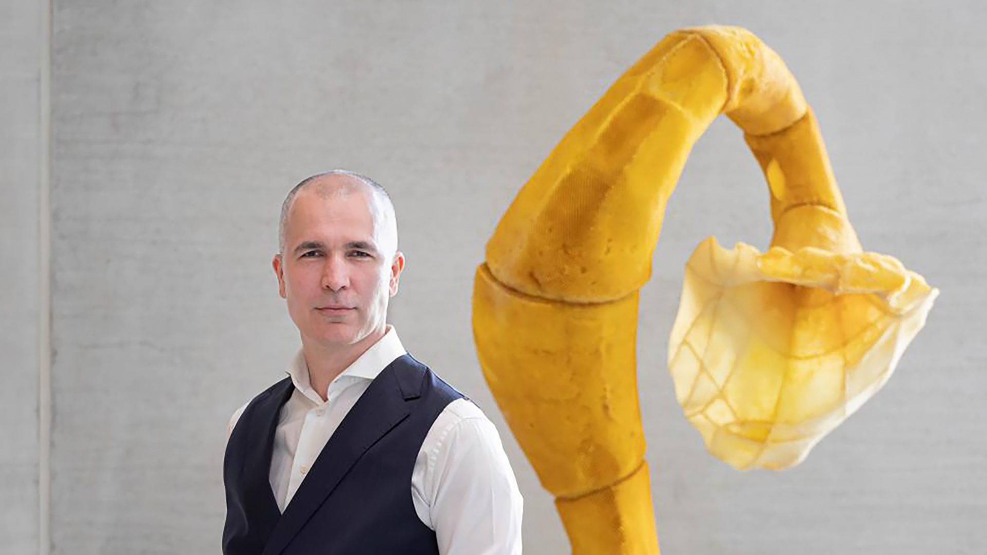 Een kale man in een donkere gilet en wit overhemd staat voor een groot, geel kunstwerk met een gebogen vorm tegen een grijze muur.