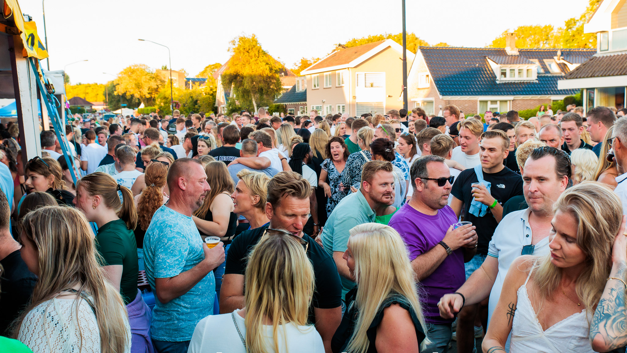 Een grote menigte mensen op straat tijdens een evenement genaamd Tour de Waard “Marlene”, op een zonnige dag met huizen op de achtergrond.