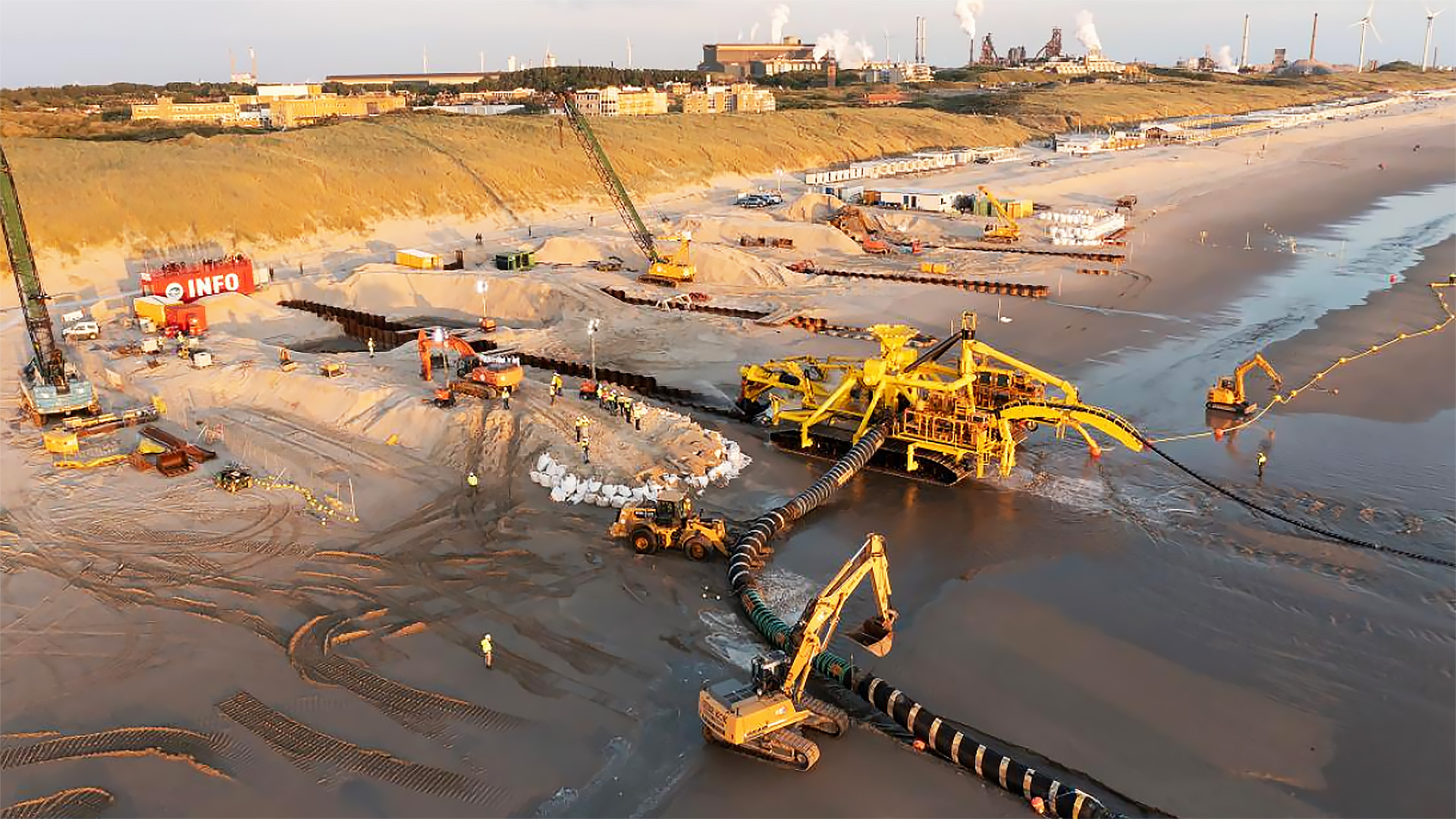 Luchtfoto van een strand met zware bouwmachines die betrokken zijn bij werkzaamheden, waaronder graafmachines en pijpleidingen. Er zijn arbeiders te zien in felgekleurde vesten, en een rode container met 'INFO' erop staat op de achtergrond bij de duinen.