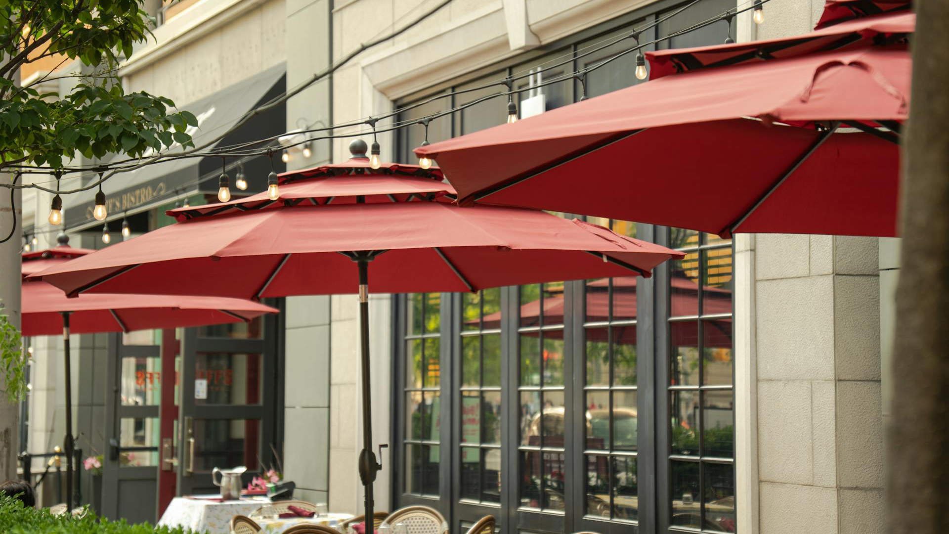 Buitenterras van een café met rode parasols en tafels, omringd door bakstenen muren en verlichting aan snoeren boven de tafels.
