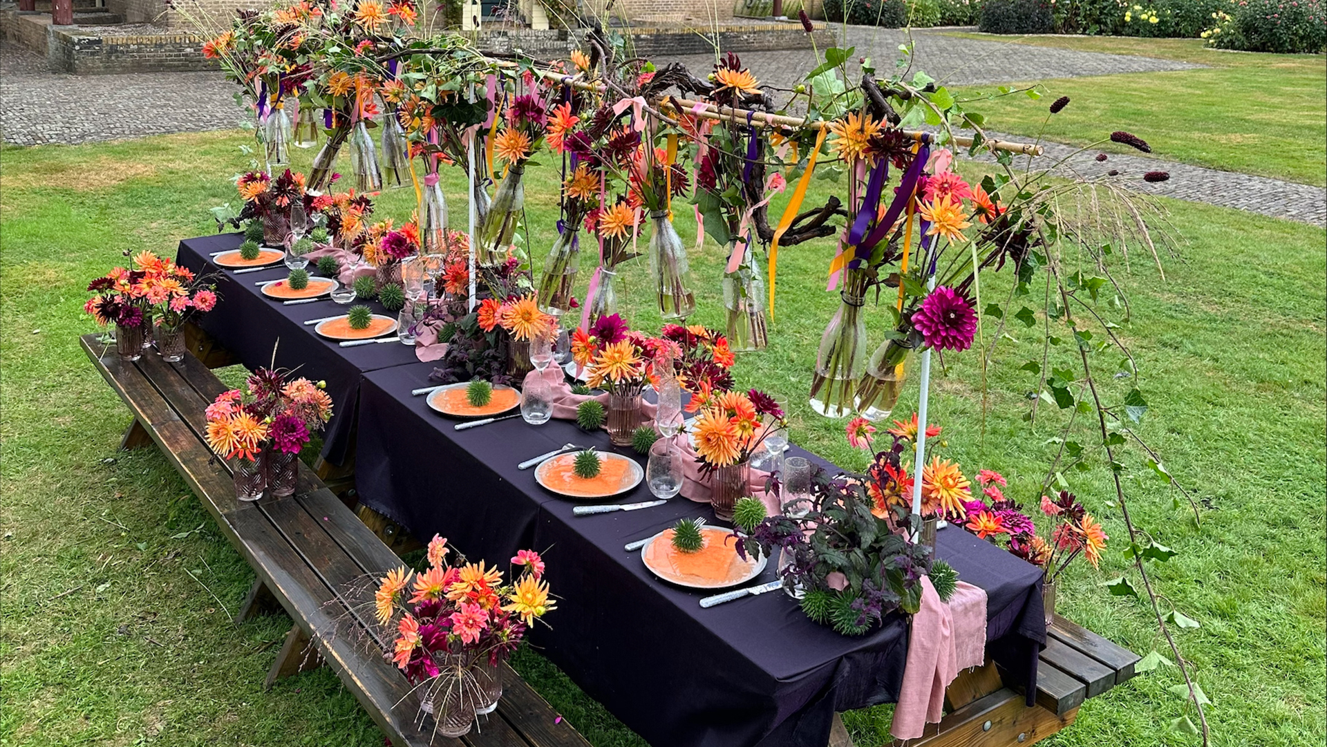 Langwerpige eettafel buiten gedekt met zwart tafelkleed, kleurrijke bloemenarrangementen en borden met oranje decoraties. Boven de tafel hangen vaasjes met bloemen, vastgebonden met linten, en op de tafel staan glazen, servetten en bestek. De omgeving is een grasveld met een bestrate achtergrond.