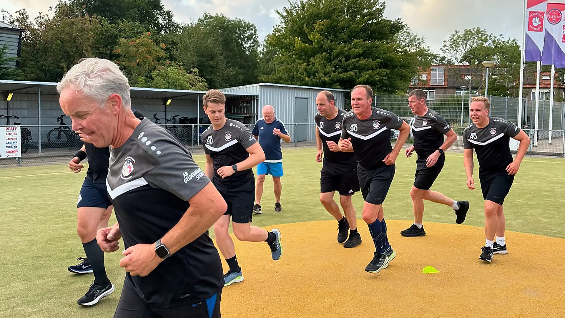 Een groep mannen in sportkleding loopt over een sportveld tijdens een trainingssessie, met enkele fietsen en een container op de achtergrond.