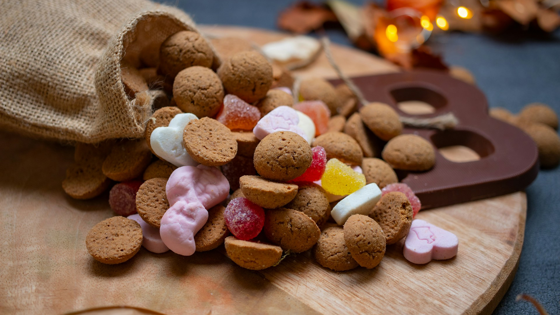Gevulde jute zak met kruidnoten en gekleurde snoepjes, naast een chocoladeletter op een houten plank.