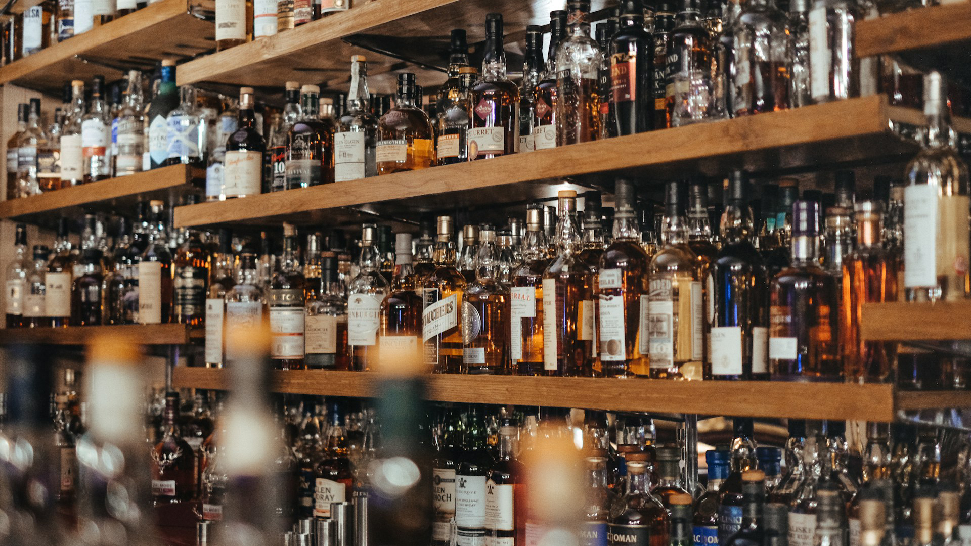 Houten planken vol met een gevarieerde collectie flessen sterke drank, waaronder whisky, in een bar of wijnkelder.