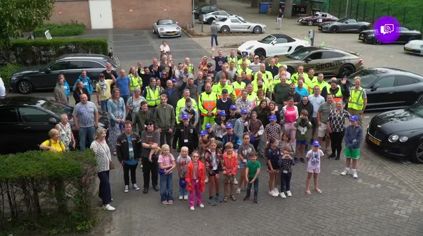 Een grote groep mensen, waaronder kinderen en volwassenen in veiligheidskleding, poseren samen op een parkeerplaats. Er staan auto's geparkeerd rondom de groep, en sommige kinderen dragen blauwe petten.