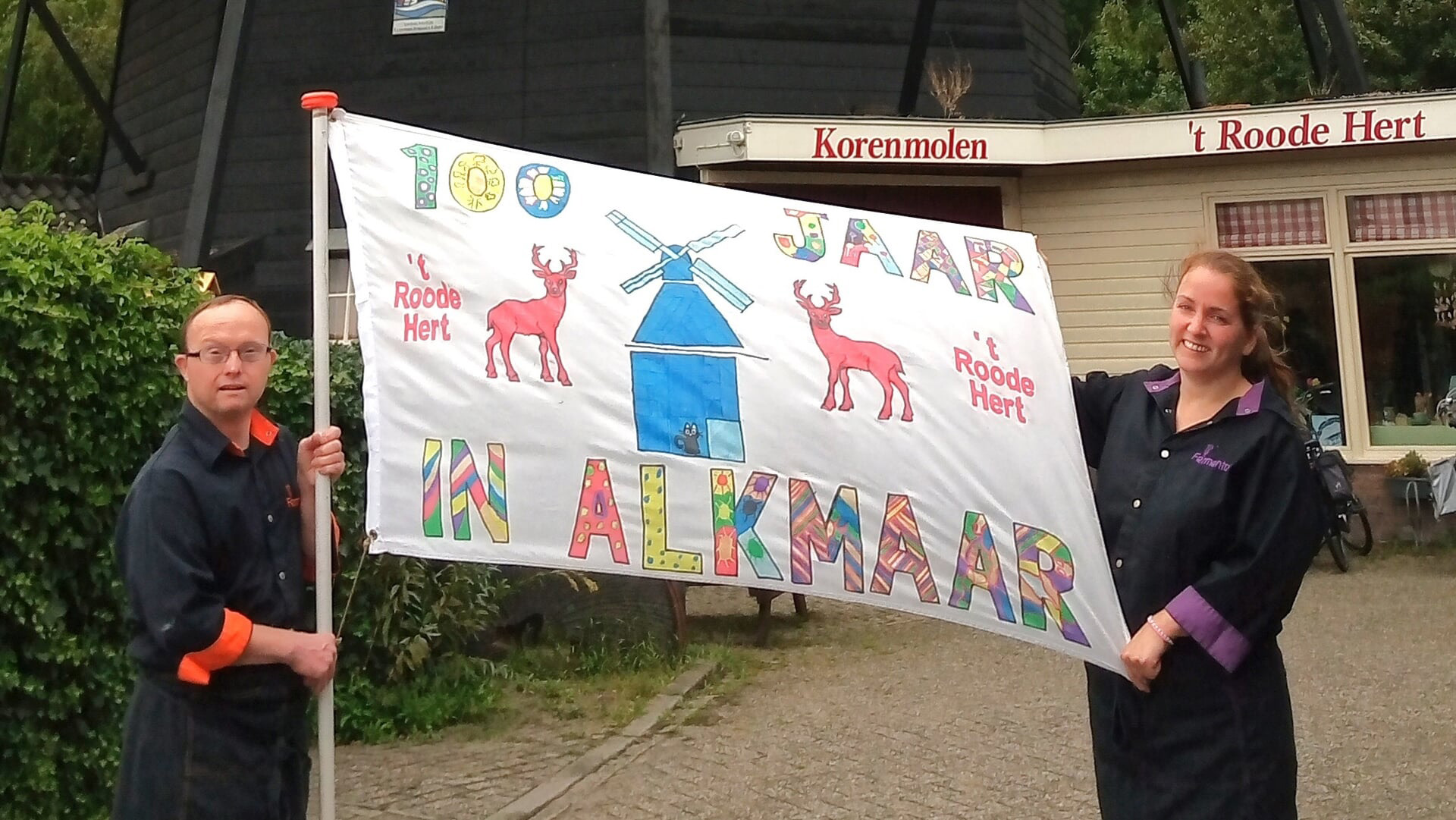 Twee mensen houden een vlag vast met de tekst '100 jaar in Alkmaar' en afbeeldingen van een molen en herten, voor gebouw 't Roode Hert.'