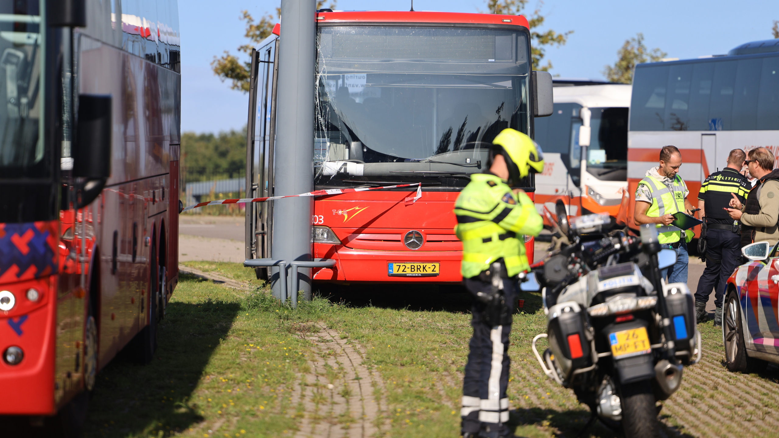 Een rode bus is tegen een lantaarnpaal gebotst, omringd door politie en verkeersveiligheidsfunctionarissen met motorvoertuigen. Er zijn meerdere politieagenten aanwezig voor onderzoek. De voorkant van de bus is beschadigd.