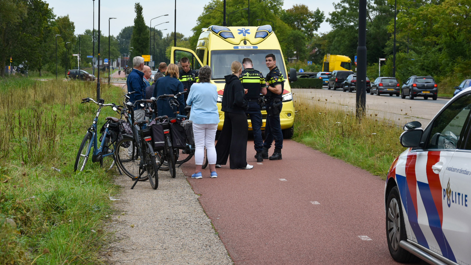 Groep mensen en politieagenten staan bij fietsen naast een ambulance op een fietspad.