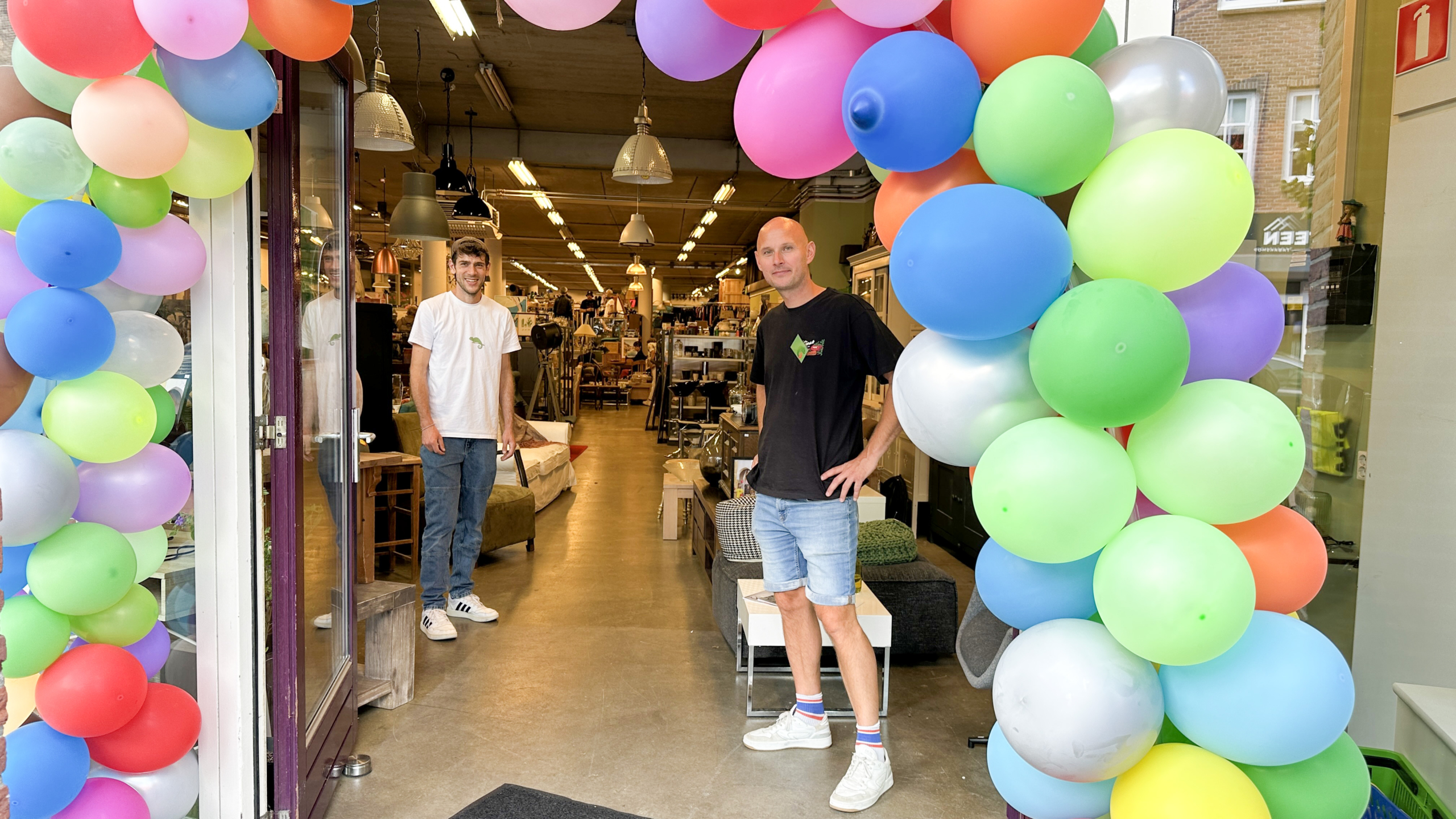 Twee mannen staan glimlachend bij de ingang van een winkel, versierd met kleurrijke ballonnen, met verschillende meubels en verlichting zichtbaar binnen.