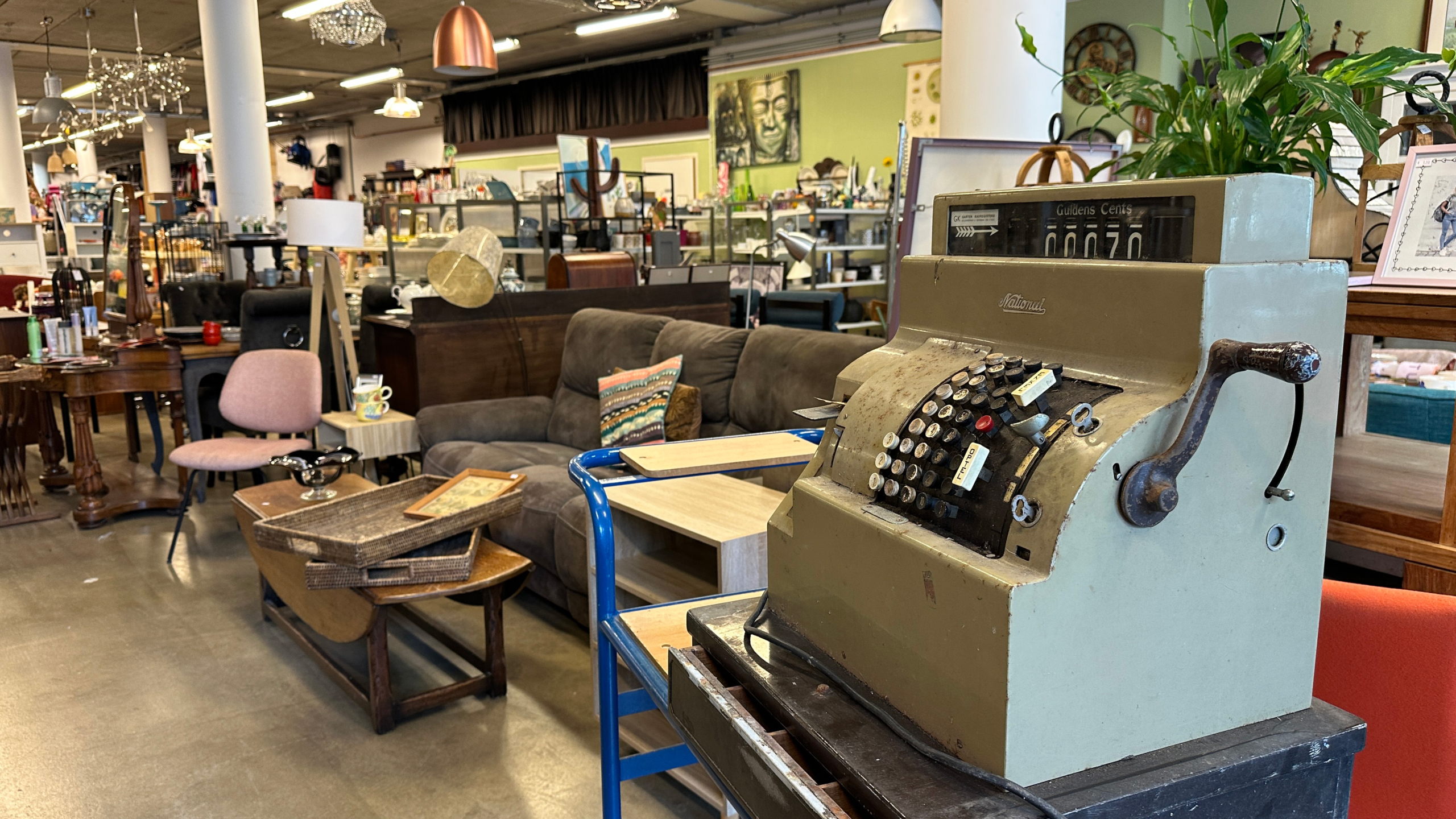 Antieke kassa met een handmatige hendel in een tweedehandswinkel vol met meubels en decoratieve items zoals stoelen, lampen en schilderijen.