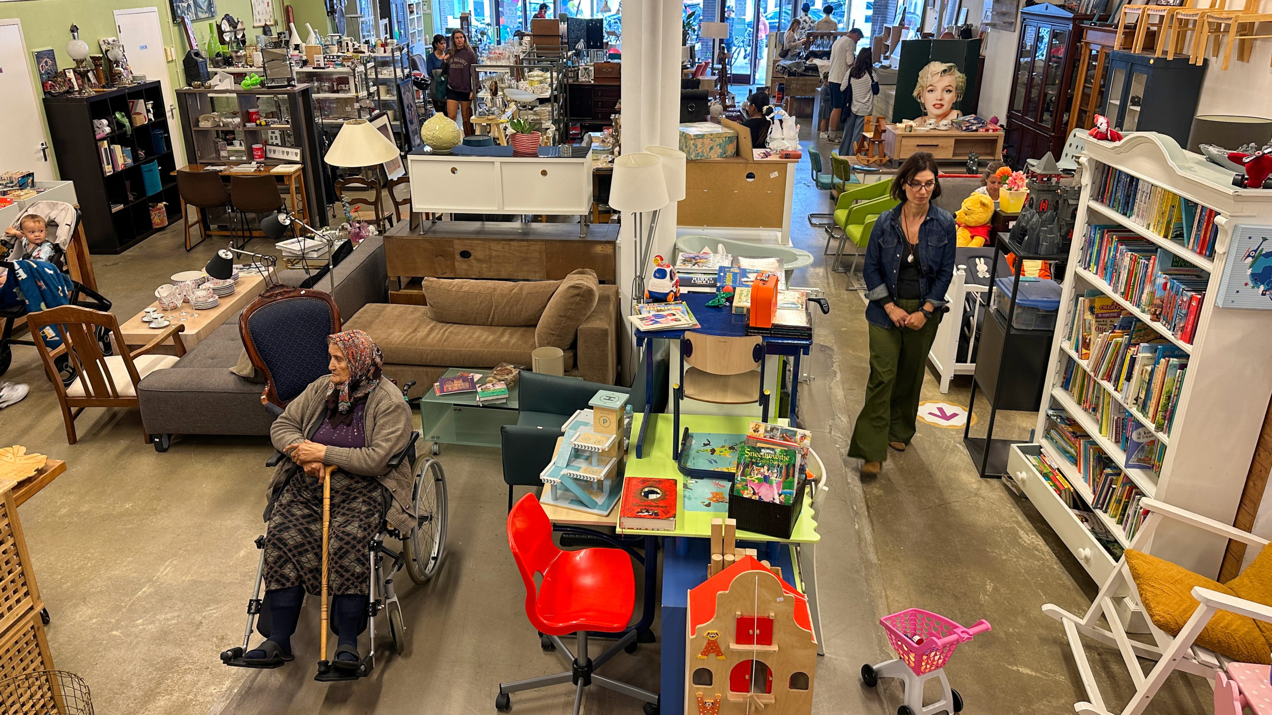 Een tweedehands winkel vol met meubels en boeken, met een oudere vrouw in een rolstoel in de voorgrond. Links zijn mensen met een kinderwagen te zien en rechts een vrouw die langs boekenplanken loopt. De winkel is gevuld met diverse objecten zoals speelgoed en decoratieve items.