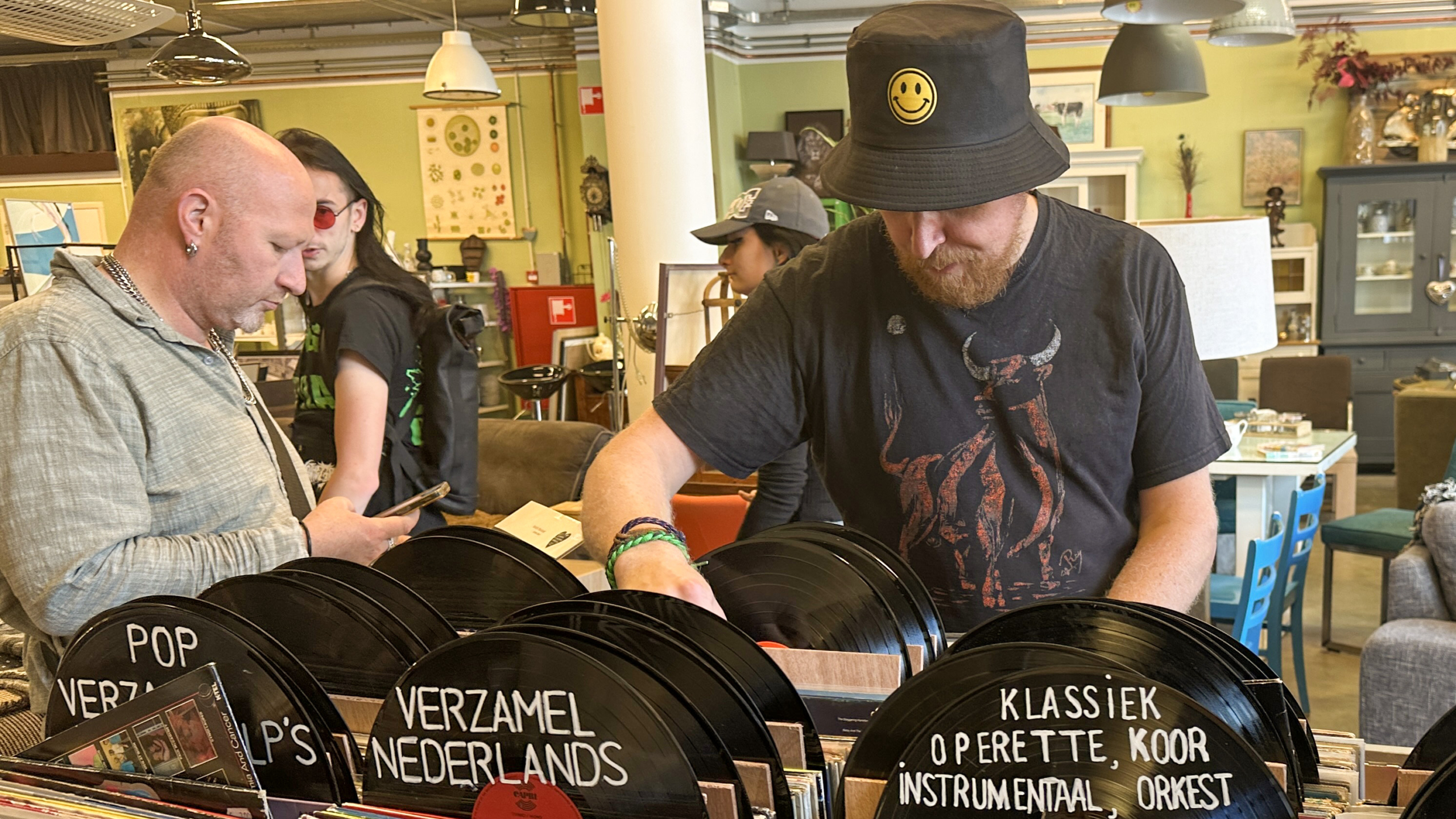 Mensen bladeren door vinylplaten in een kringloopwinkel, waaronder een man met een hoed met een smiley-embleem.