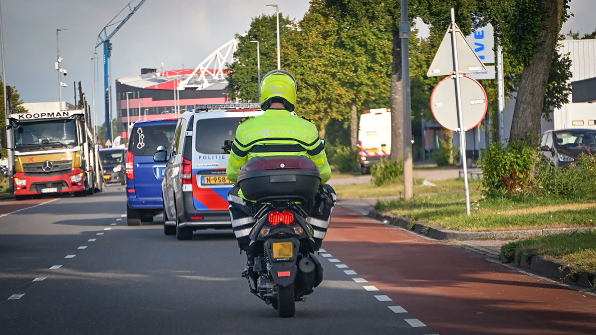 Een politieagent op een motor rijdt op een weg achter een politiebusje, met een vrachtwagen links en enkele bomen aan de kant van de weg.