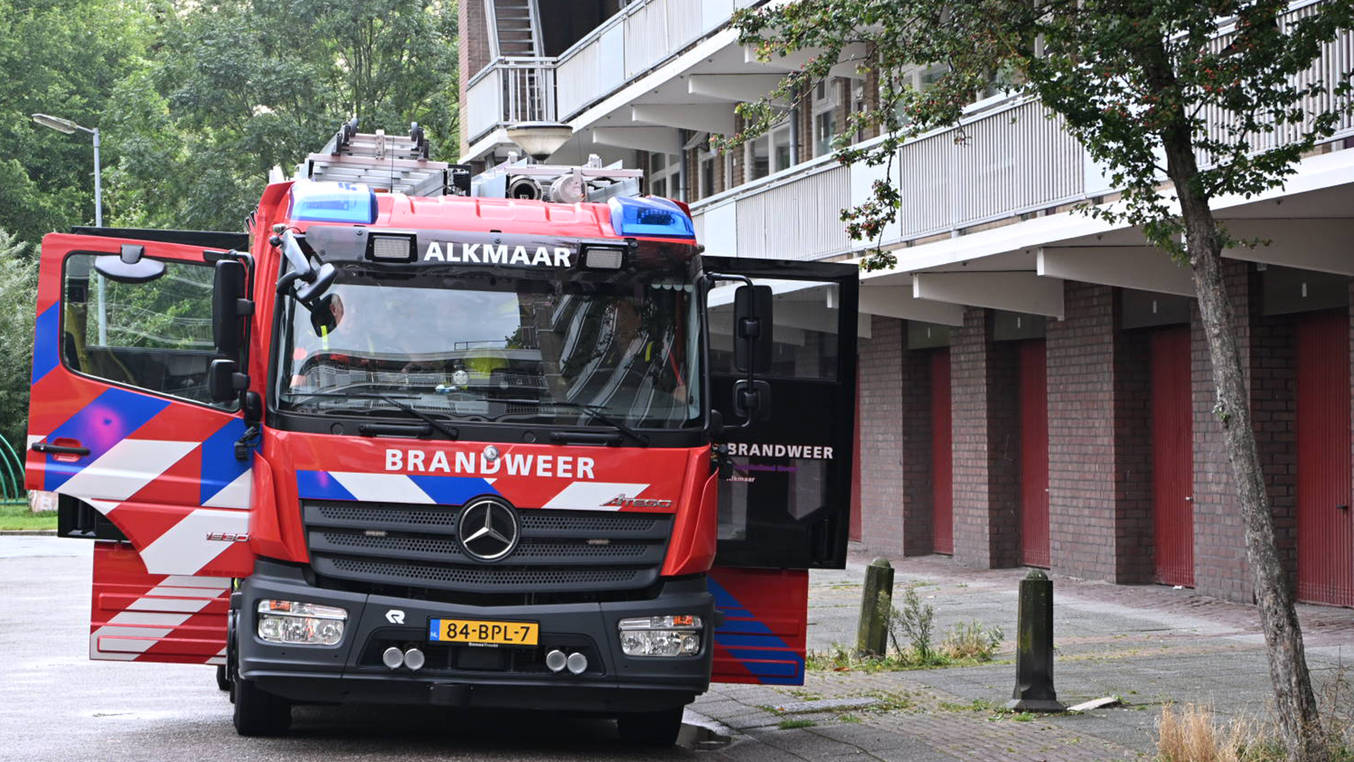 Een rode brandweerwagen van de brandweer Alkmaar met geopende portieren, geparkeerd voor een gebouw met bakstenen muren en balkons.