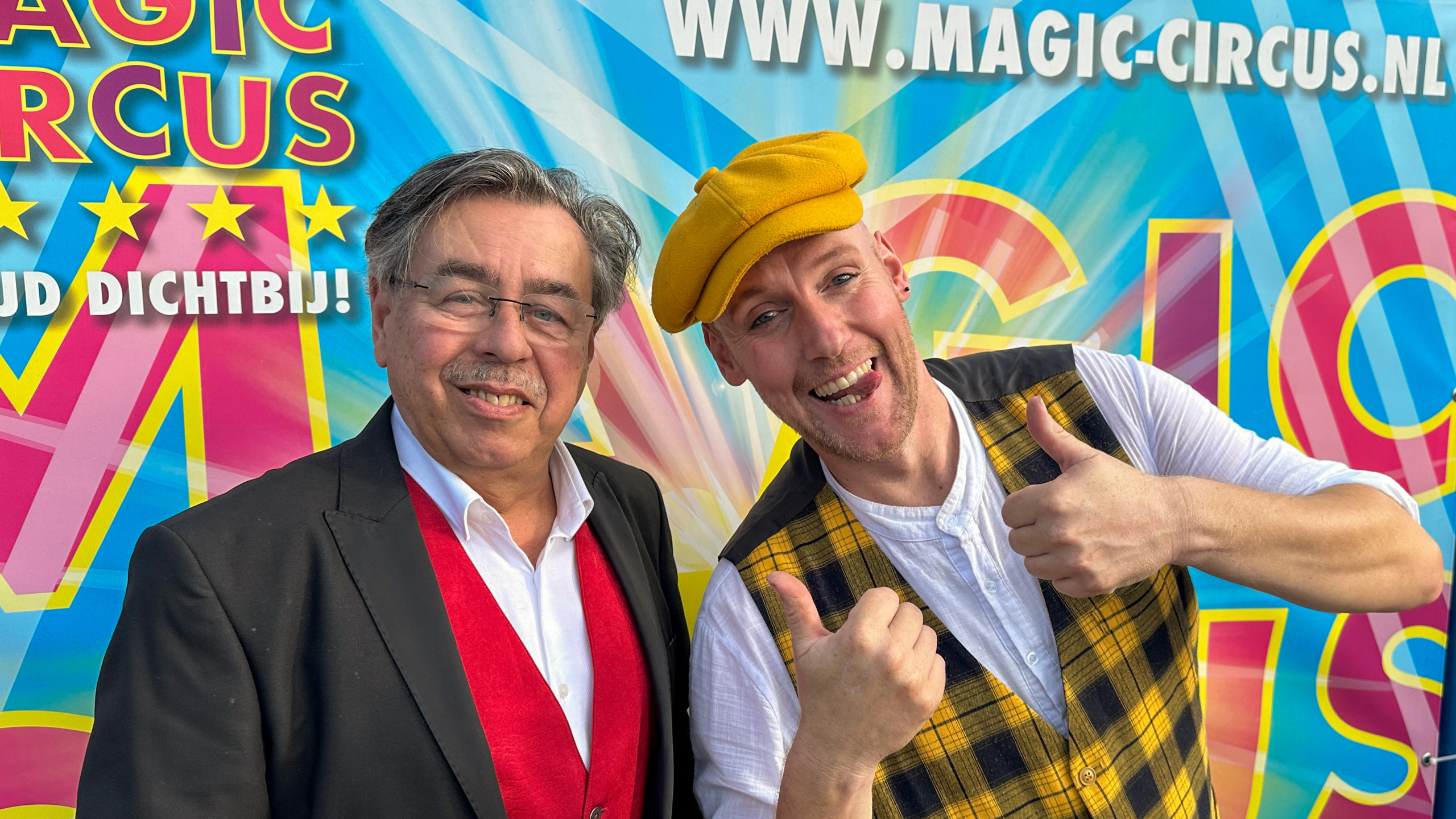Twee mannen poseren voor een kleurrijke poster van het Magic Circus; de man links draagt een zwart colbert met een rood vest en een witte overhemd, en de man rechts draagt een geel petje en een geel-zwart geruite vest terwijl hij zijn duim omhoogsteekt en glimlacht.