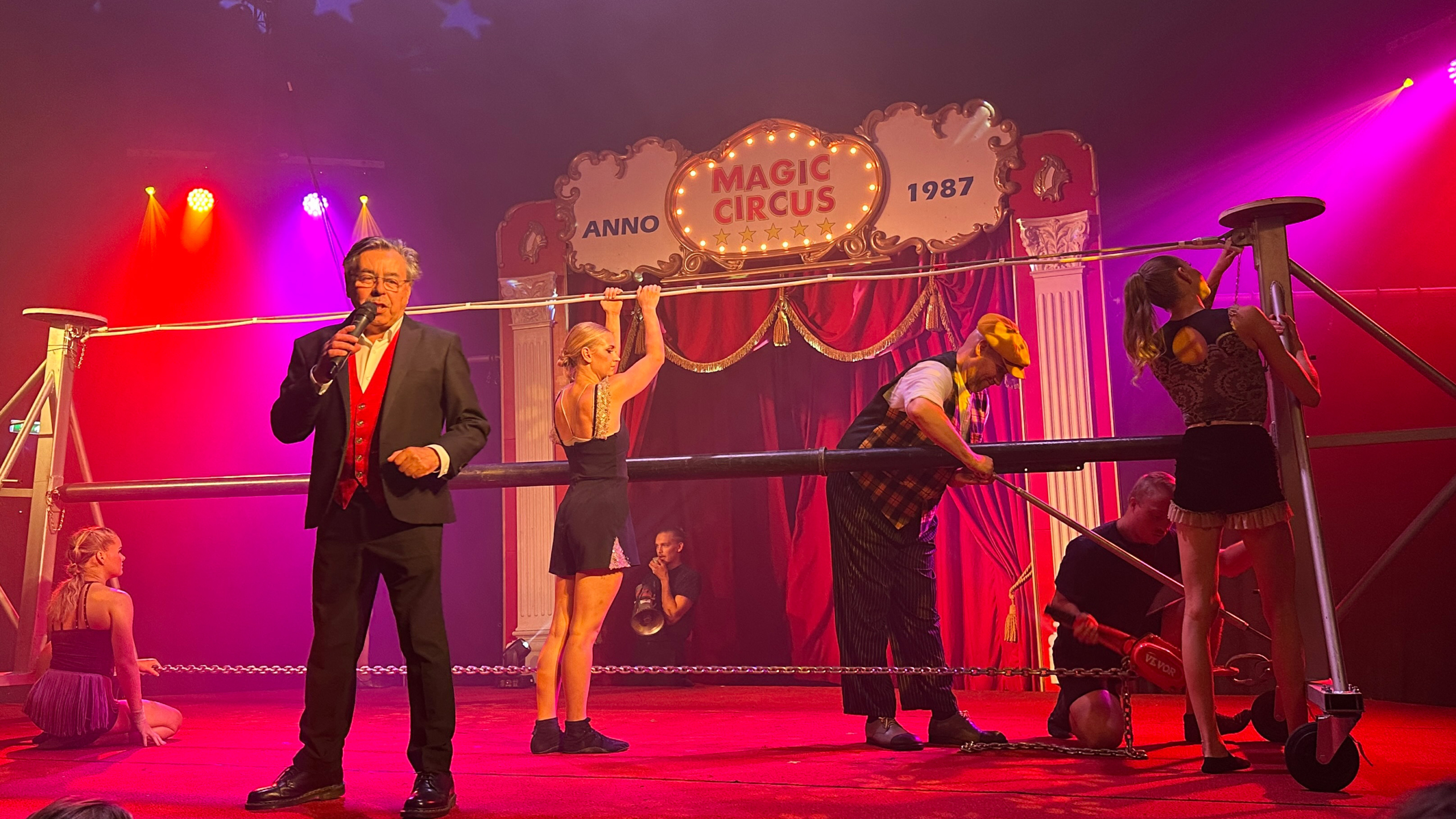 Circusvoorstelling met een man in een zwart pak en rode gilet die een microfoon vasthoudt, omgeven door artiesten die een hoge trapeze installeren op een podium met een rood gordijn en een bord dat "Magic Circus" aangeeft.