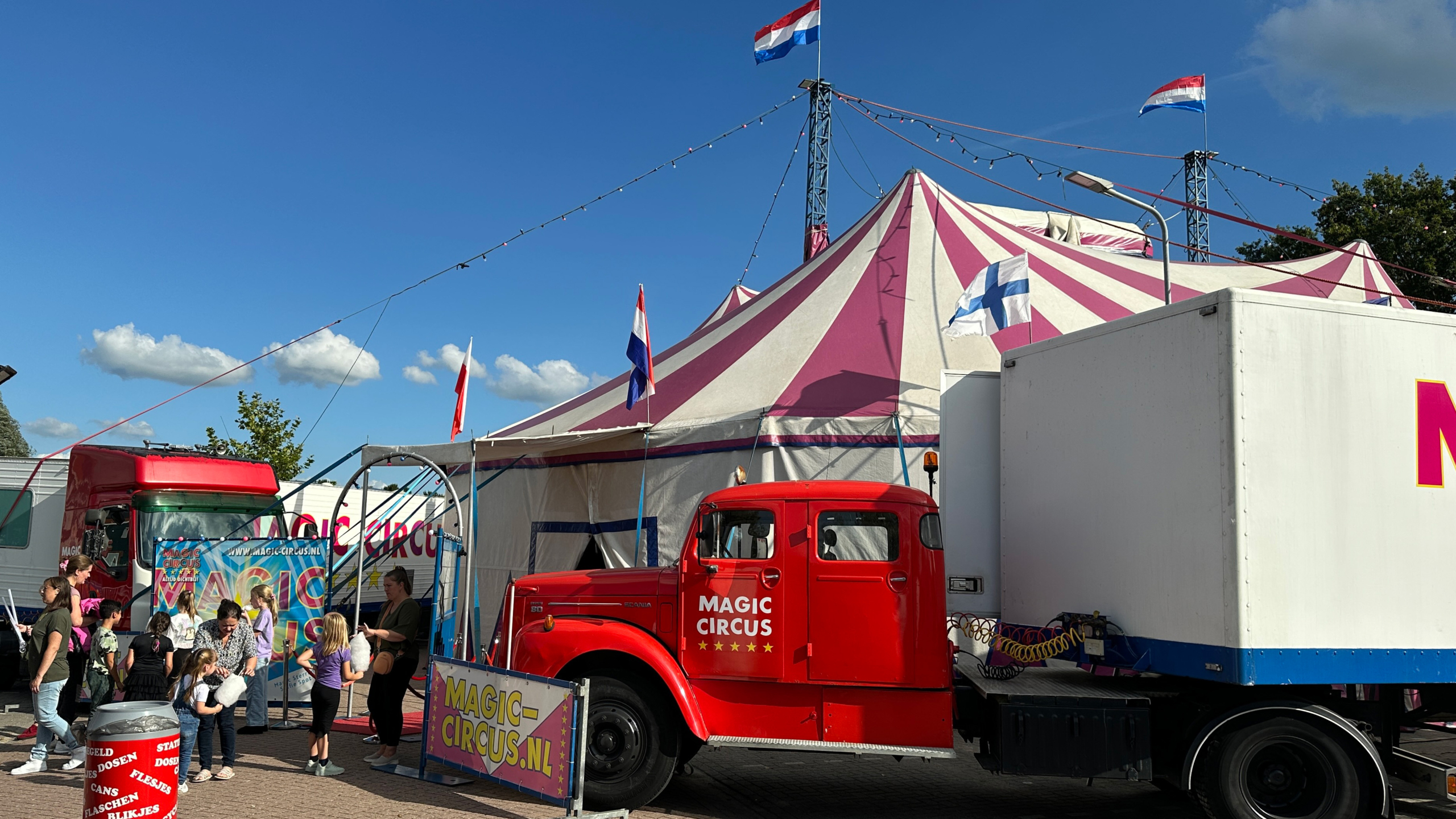 Circustent met rode vrachtwagen en mensen die in de rij staan op een zonnige dag, met Nederlandse en andere vlaggen op de achtergrond.