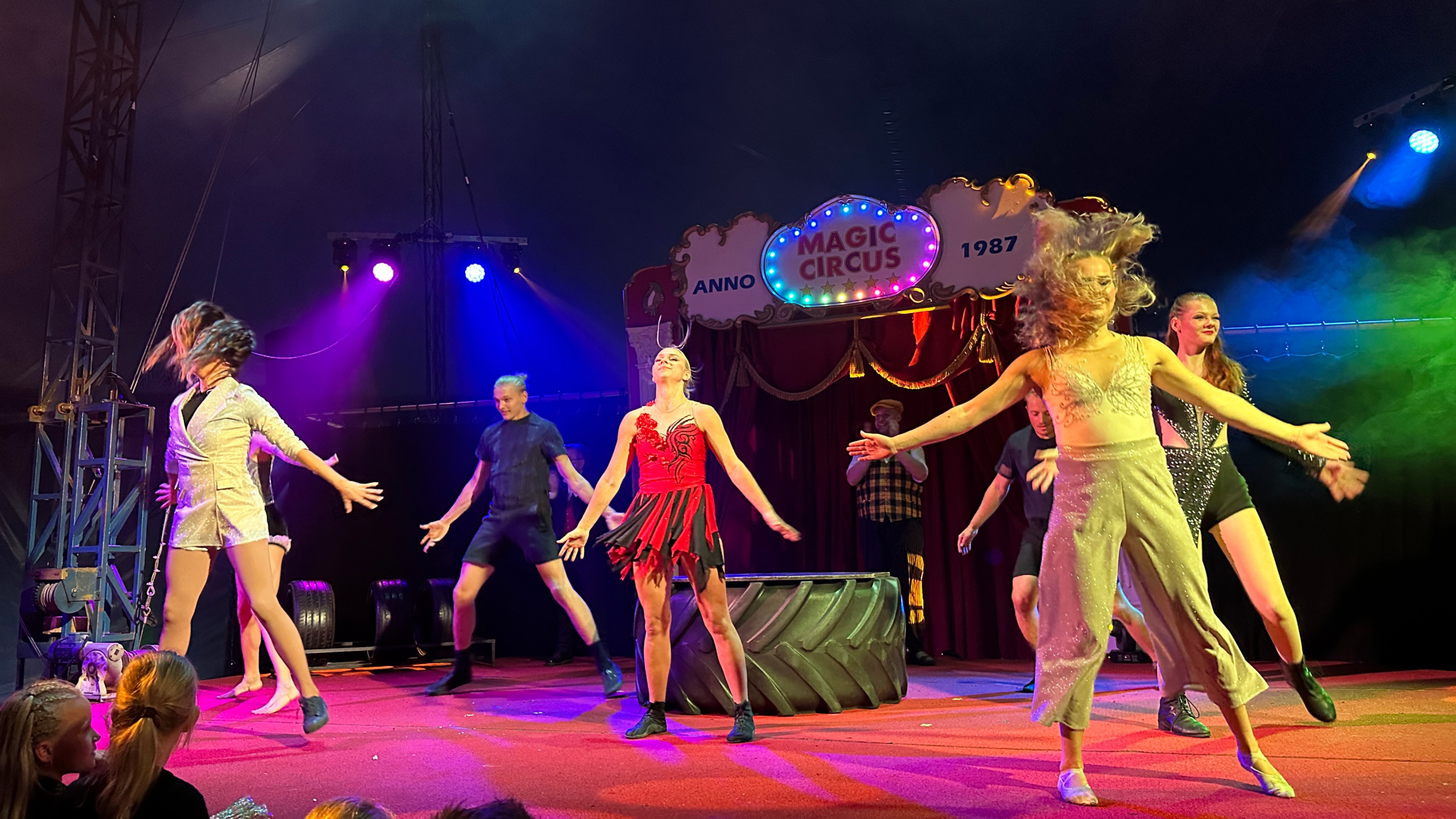 Een groep artiesten voert een energieke dansroutine op in het Magic Circus, met felgekleurde verlichting op de achtergrond en publiek op de voorgrond.