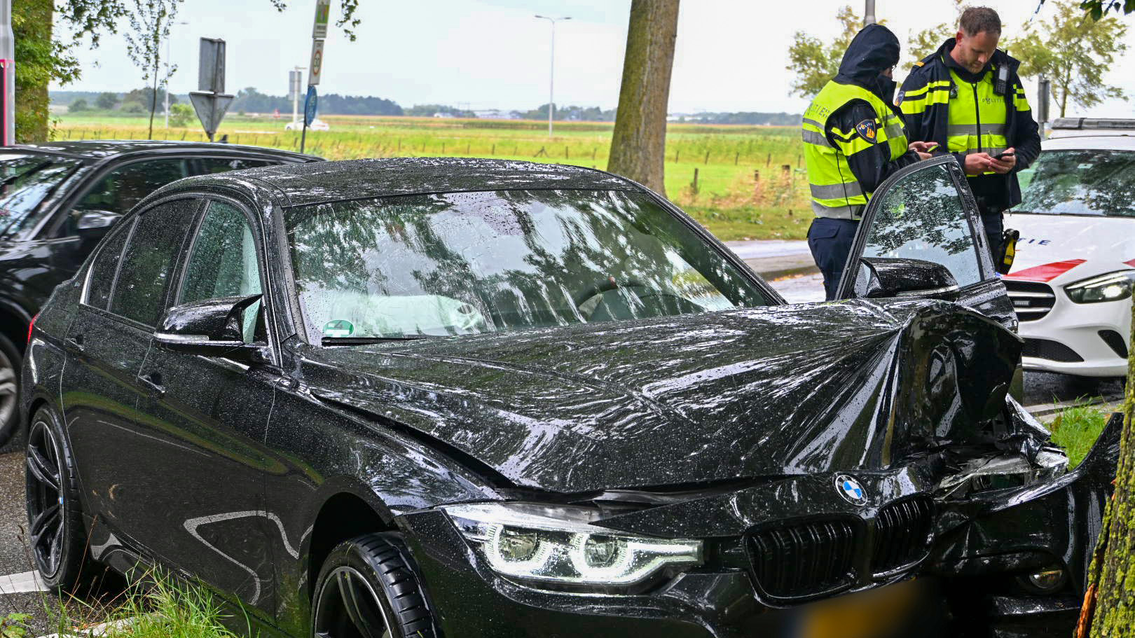 Een beschadigde zwarte BMW met een ingedeukte voorkant na een botsing tegen een boom, met op de achtergrond twee politieagenten die op hun smartphone kijken en een politieauto geparkeerd naast de weg.