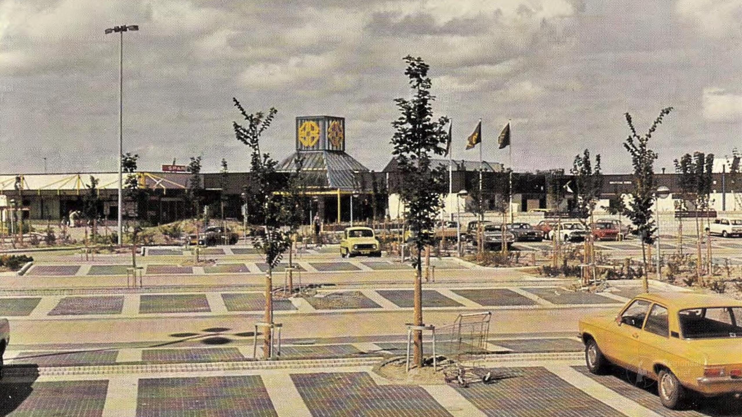 Parkeerplaats voor een supermarkt met een geel logo, omgeven door geparkeerde auto's en jonge bomen.