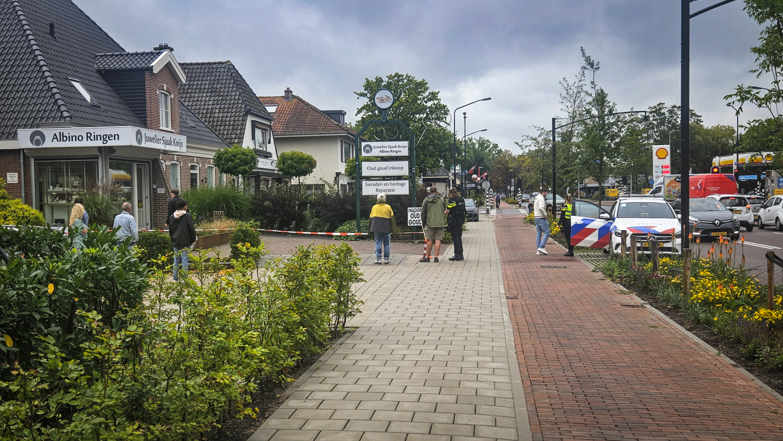 Straatbeeld in een winkelgebied met mensen die langs een juwelier lopen, een politieauto geparkeerd langs de stoep en diverse winkels aan beide zijden. Op de achtergrond zijn verkeersborden en bomen zichtbaar.