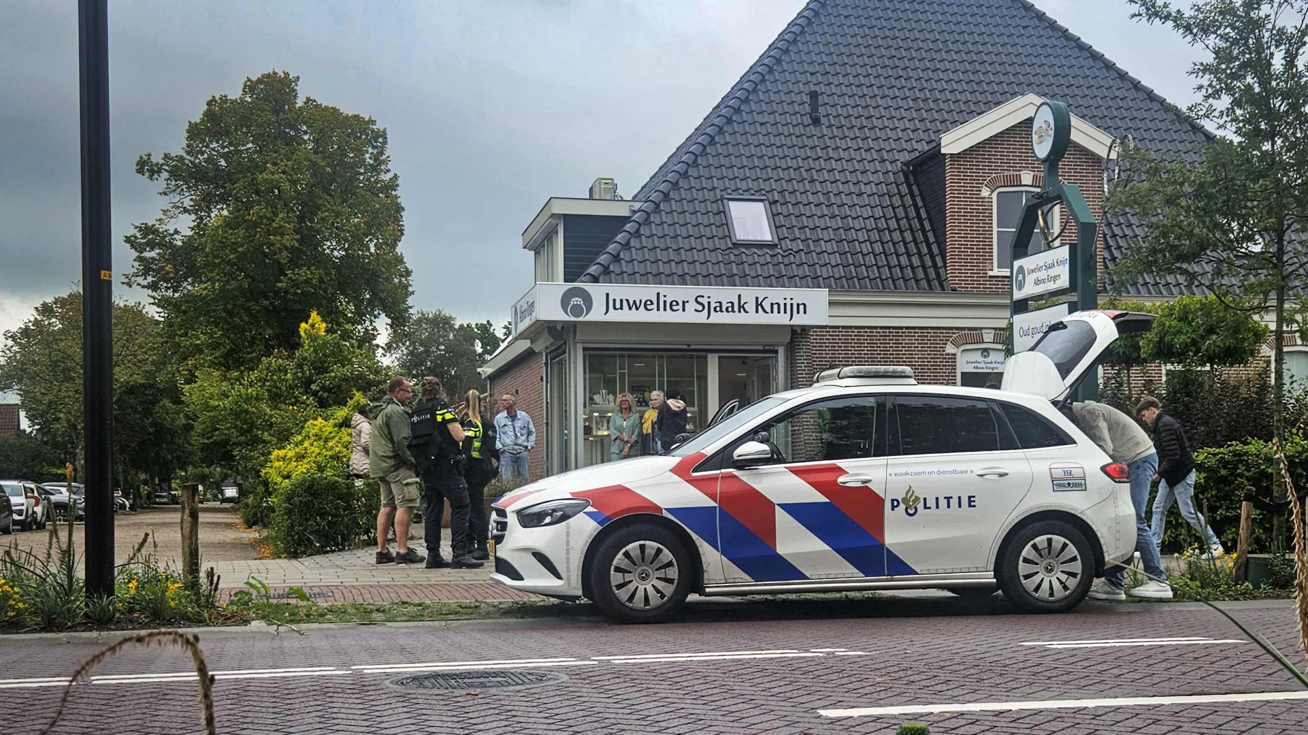 Een foto van een politieauto geparkeerd voor Juwelier Sjaak Knijn, met enkele agenten en mensen buiten de winkel in gesprek. Op de achtergrond is een bakstenen gebouw met een schuin dak en groenblijvende planten zichtbaar. Het is bewolkt weer.