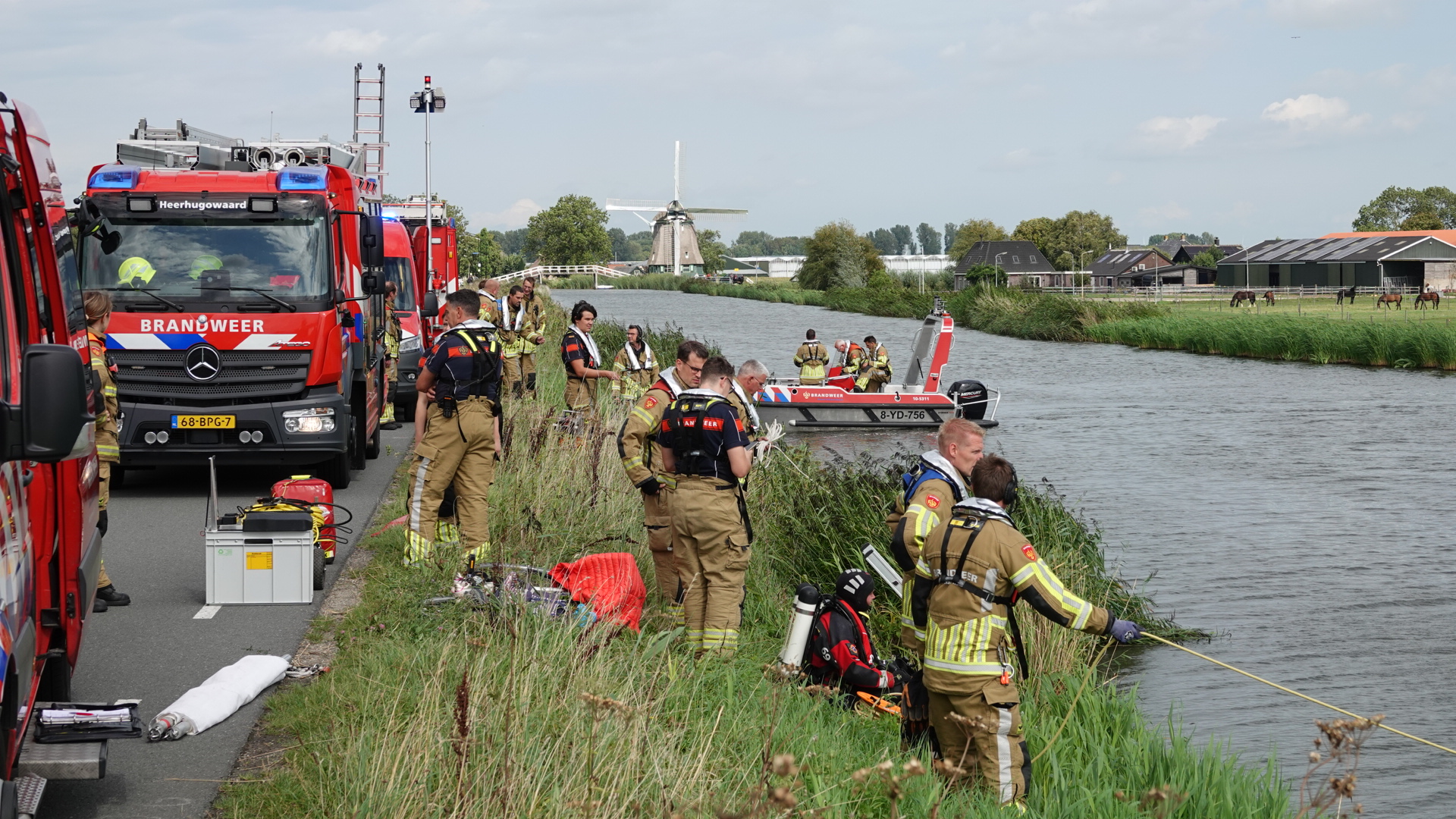 Brandweerlieden in uniformen werken langs de oever van een kanaal met enkele brandweerwagens en een reddingsboot. Op de achtergrond is een traditionele Nederlandse windmolen zichtbaar naast een weiland met enkele paarden.