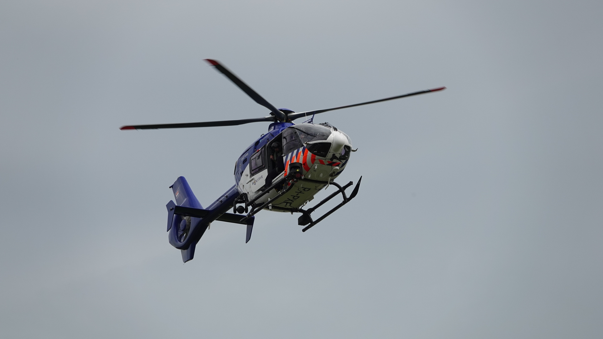 Een politiehelikopter in de lucht met draaiende rotorbladen tegen een grijze hemel.