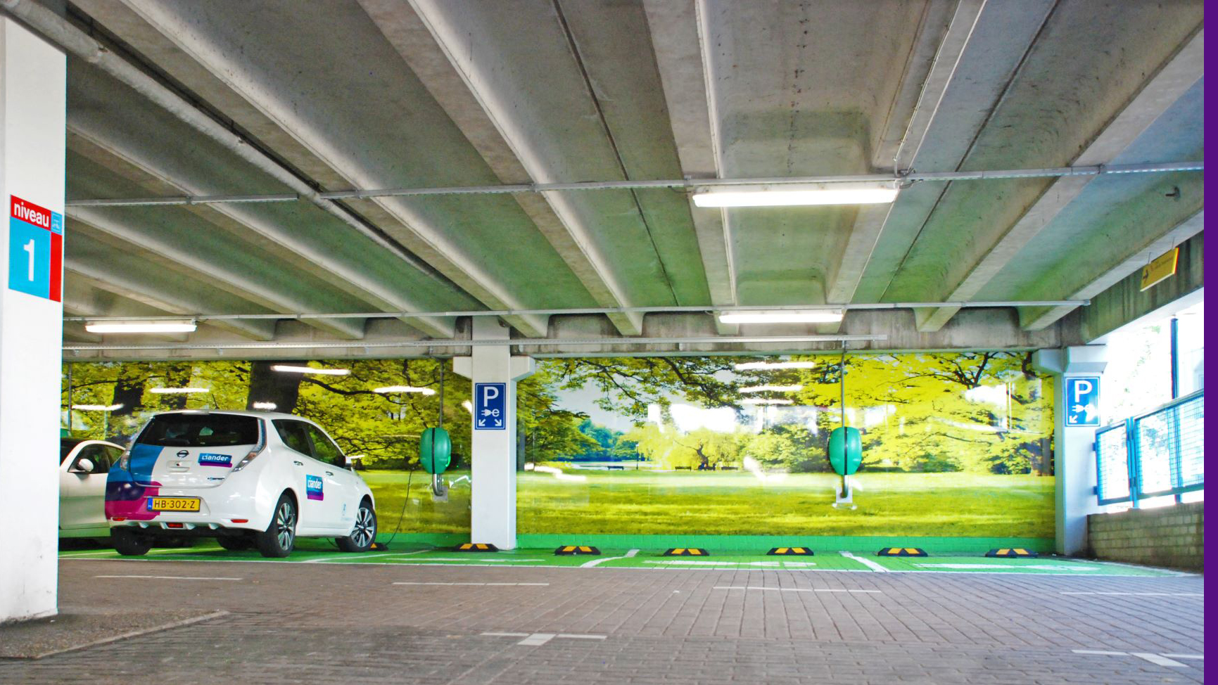 Een parkeergarage met een wit elektrische auto geparkeerd naast een wand bedrukt met een groen natuurtafereel, voorzien van oplaadpalen en bordjes voor elektrische voertuigen op niveau 1.