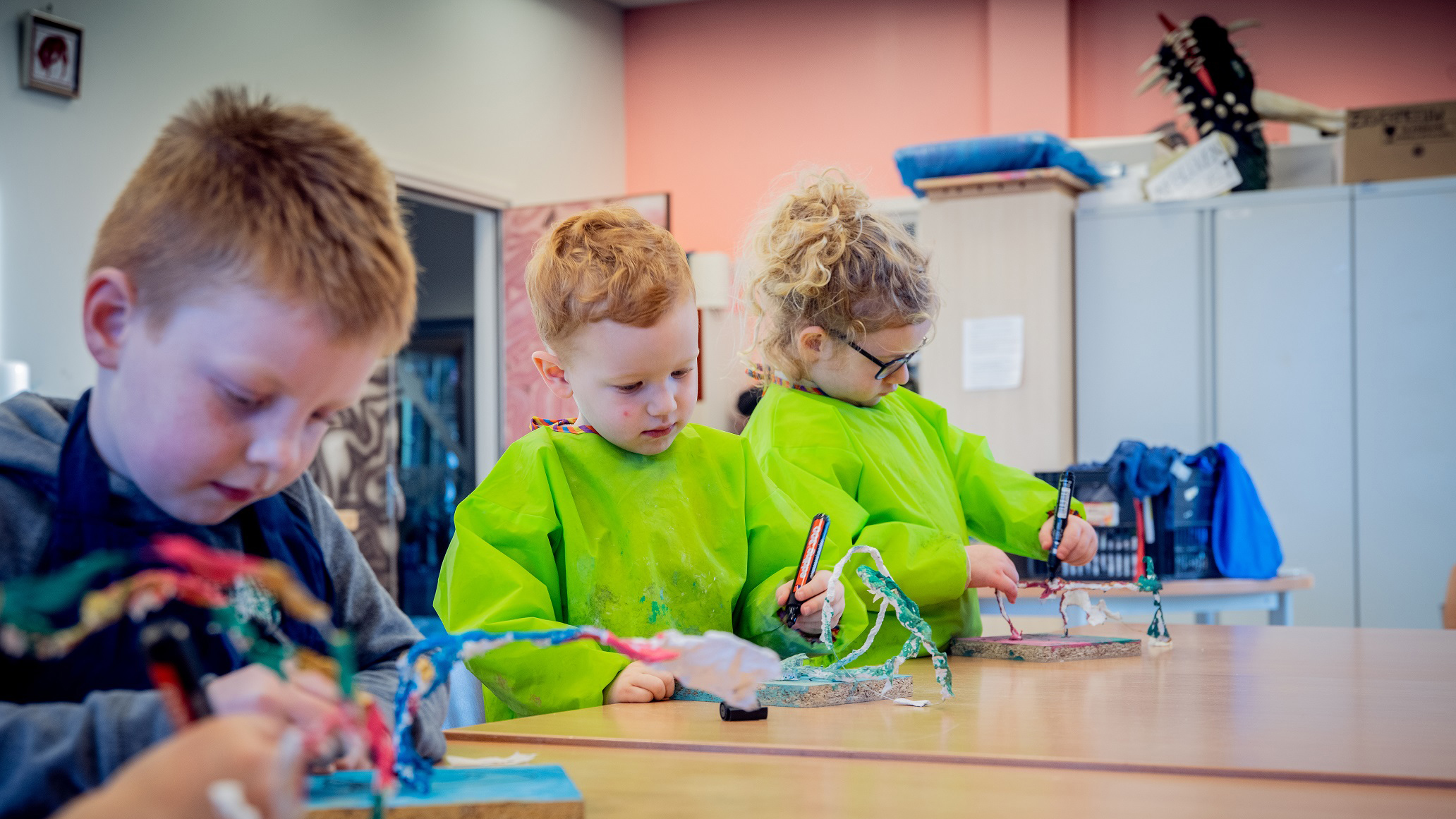 Drie kinderen in groene schorten zijn bezig met knutselen aan een tafel in een klaslokaal.