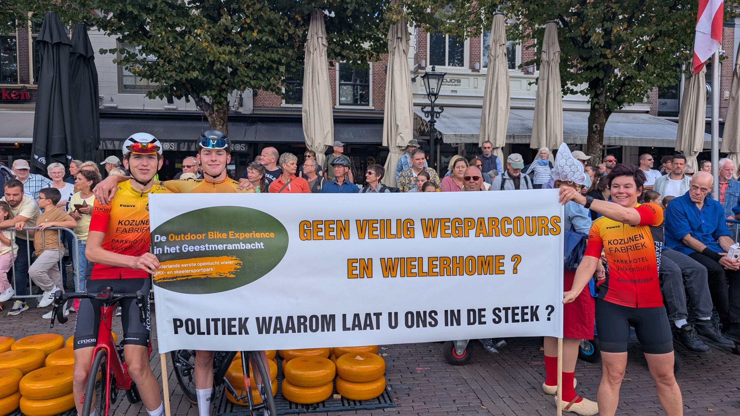 Twee wielrenners en een vrouw in wielerkleding houden een spandoek vast op een plein met toeschouwers. Op het spandoek staat een boodschap die oproept tot veilige wielerfaciliteiten. Op de achtergrond kijken mensen toe bij een evenement.