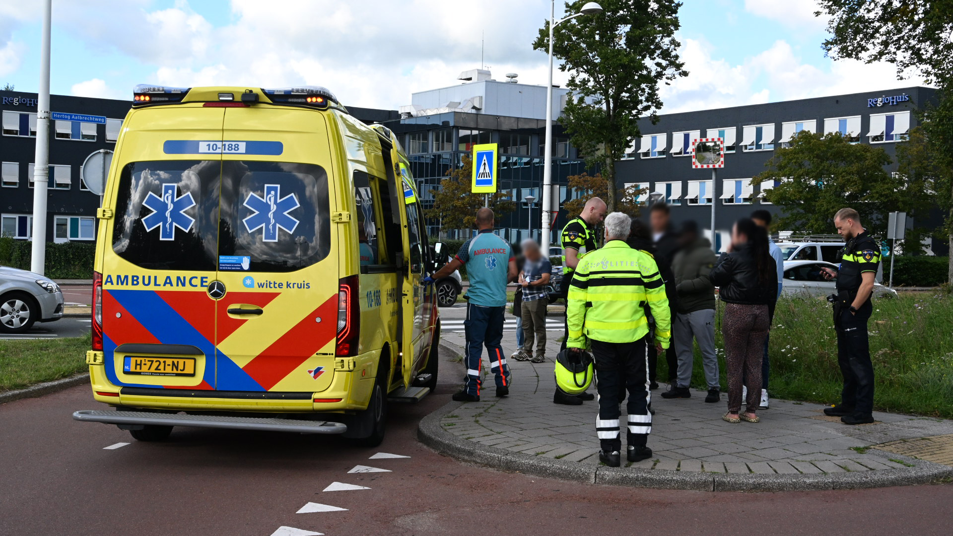 Ambulance en politie staan bij een rotonde in een stedelijke omgeving. Er is een groep mensen, waaronder een politieagent en iemand in een ambulance-uniform, in gesprek naast de ambulance.