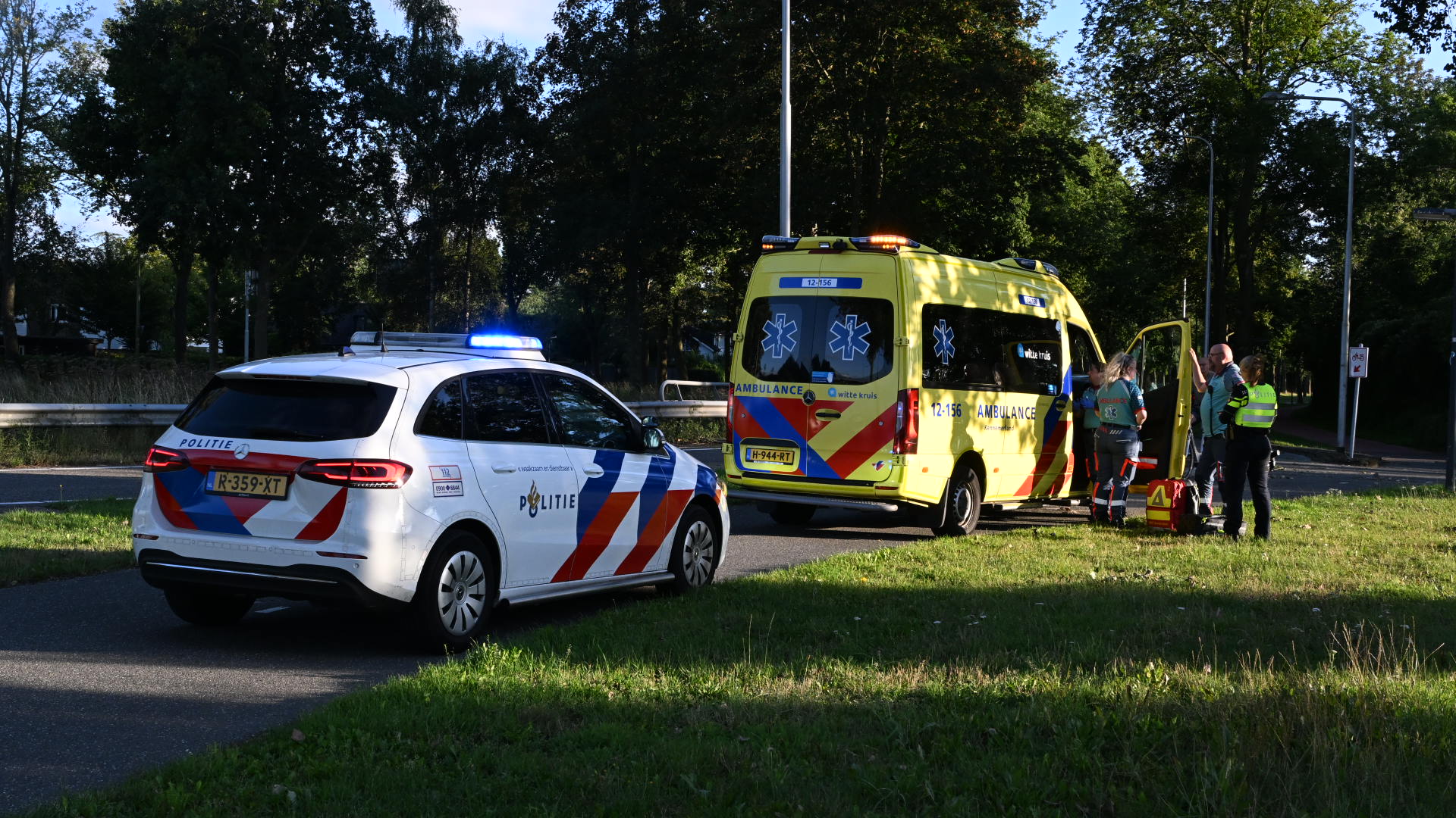 Een politieauto en een ambulance staan geparkeerd langs de weg in een groene omgeving; politieagenten en medisch personeel staan naast de ambulance in overleg.