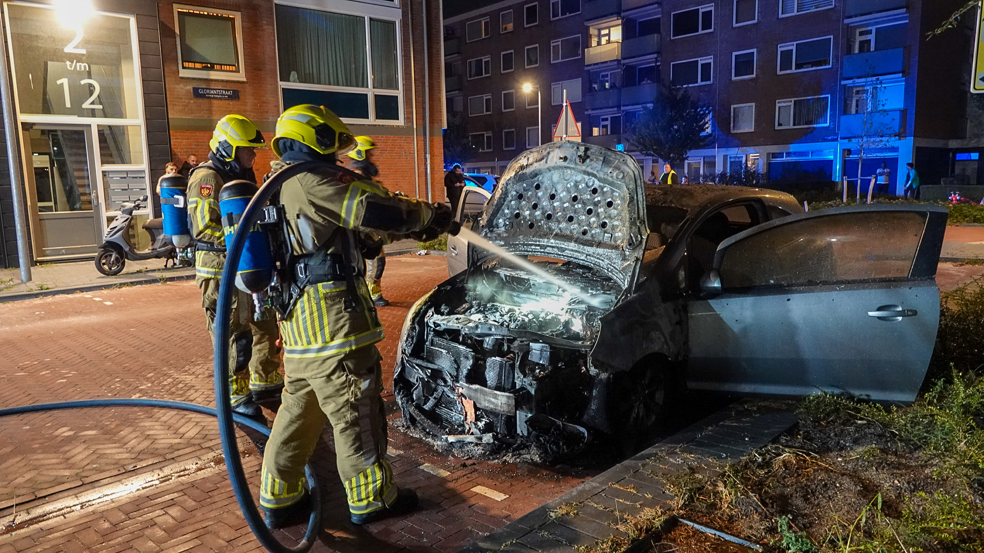 Brandweerlieden blussen 's nachts een brandende auto met open motorkap voor een flatgebouw.
