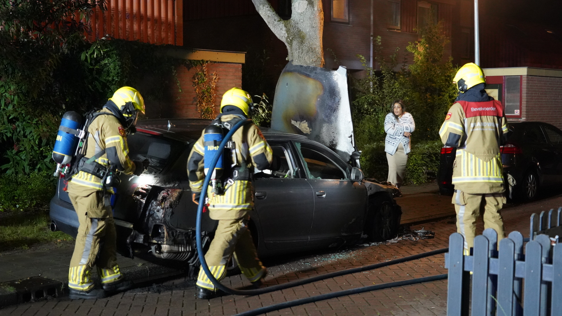 Twee brandweerlieden in gele helmen en uniformen zijn bezig met een gebluste auto naast een boom, terwijl een vrouw met een witte trui in de achtergrond toekijkt.
