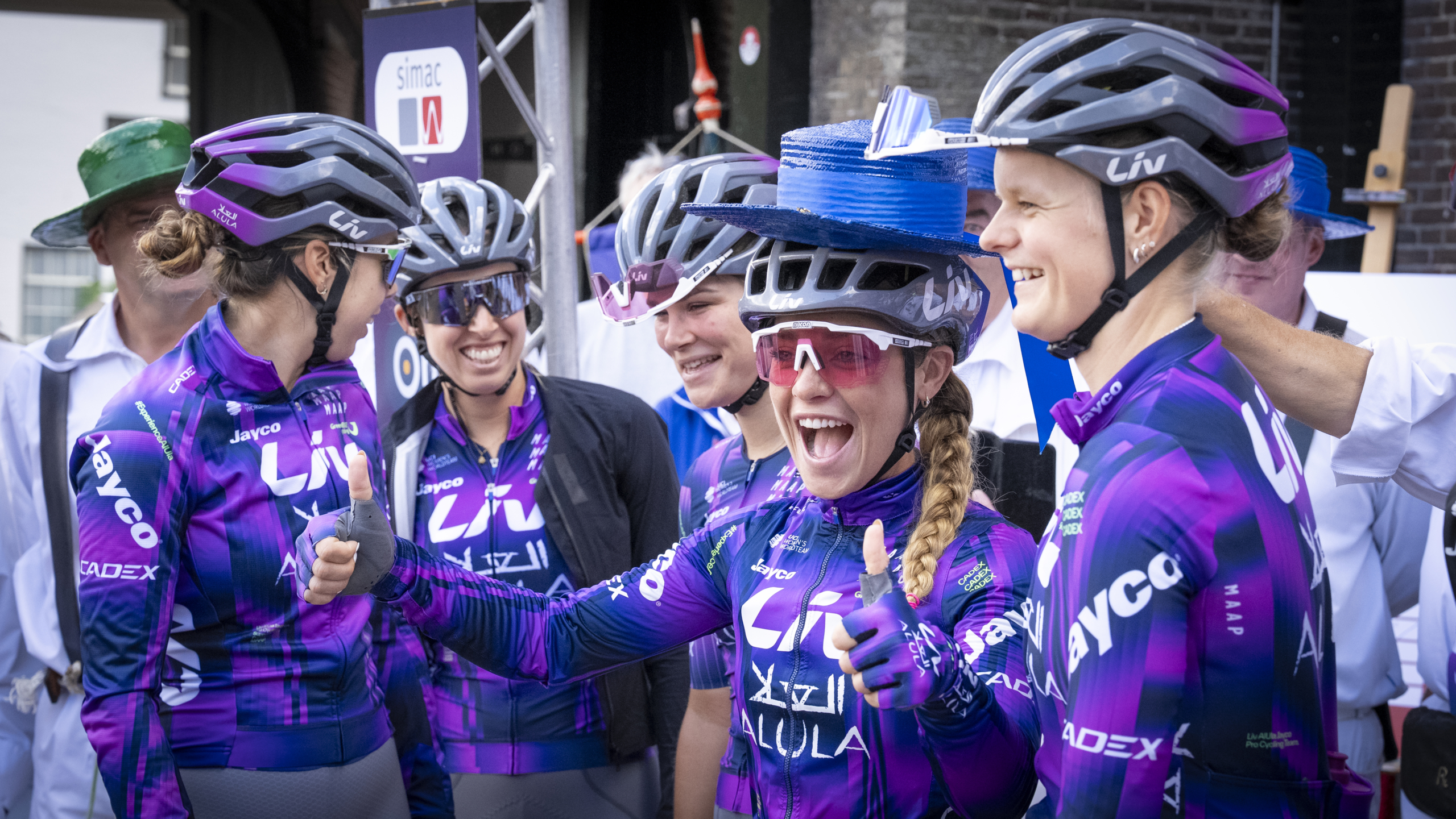 Groep vrouwelijke wielrenners in paarse Liv Alula outfits en helmen, lachen enthousiast terwijl één van hen een stapel blauwe hoeden op haar helm balanceert.