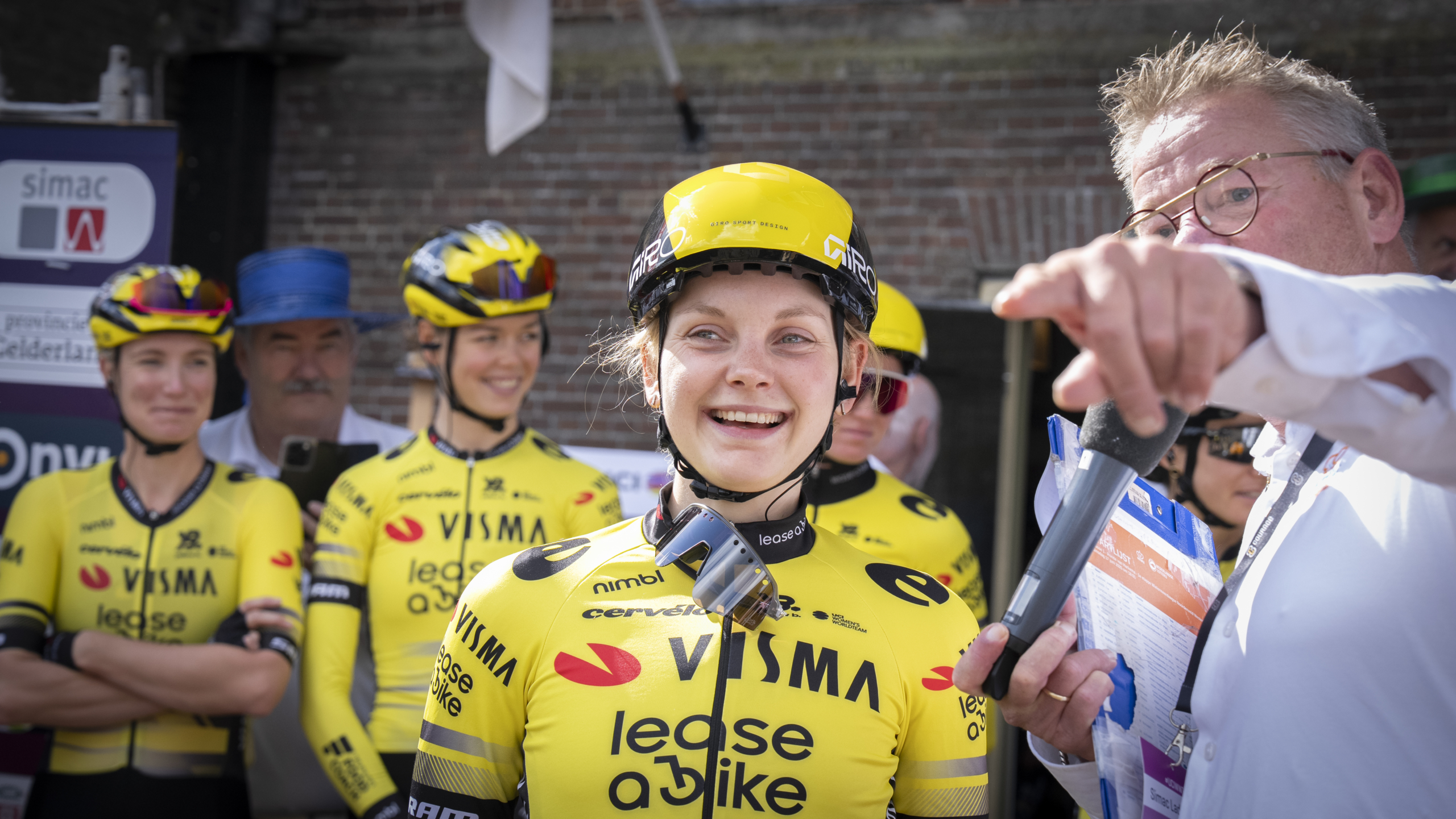 Vrouwelijke wielrenners in gele Visma Lease a Bike shirts staan lachend terwijl een man met een microfoon naar iets wijst.